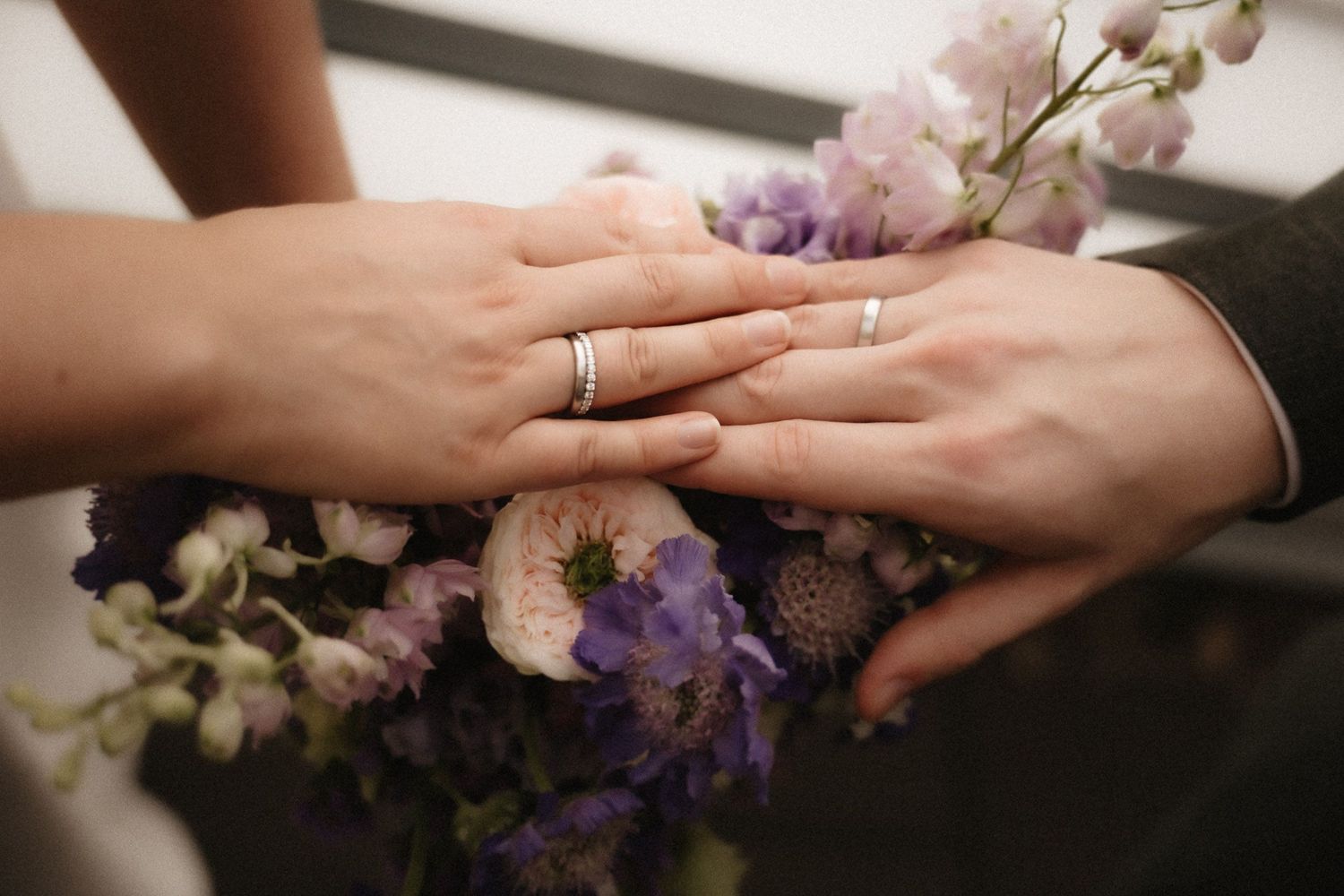 Two hands wearing wedding rings rest together on a surface with delicate pink and purple flowers.