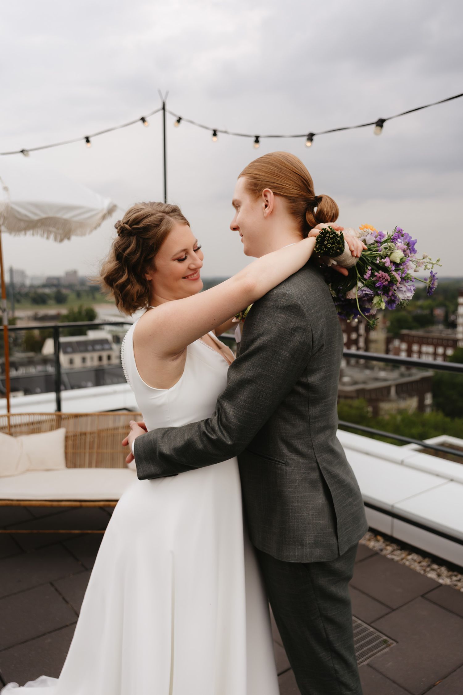 A couple in wedding attire dances and embraces on a rooftop terrace with string lights and city views in the background.