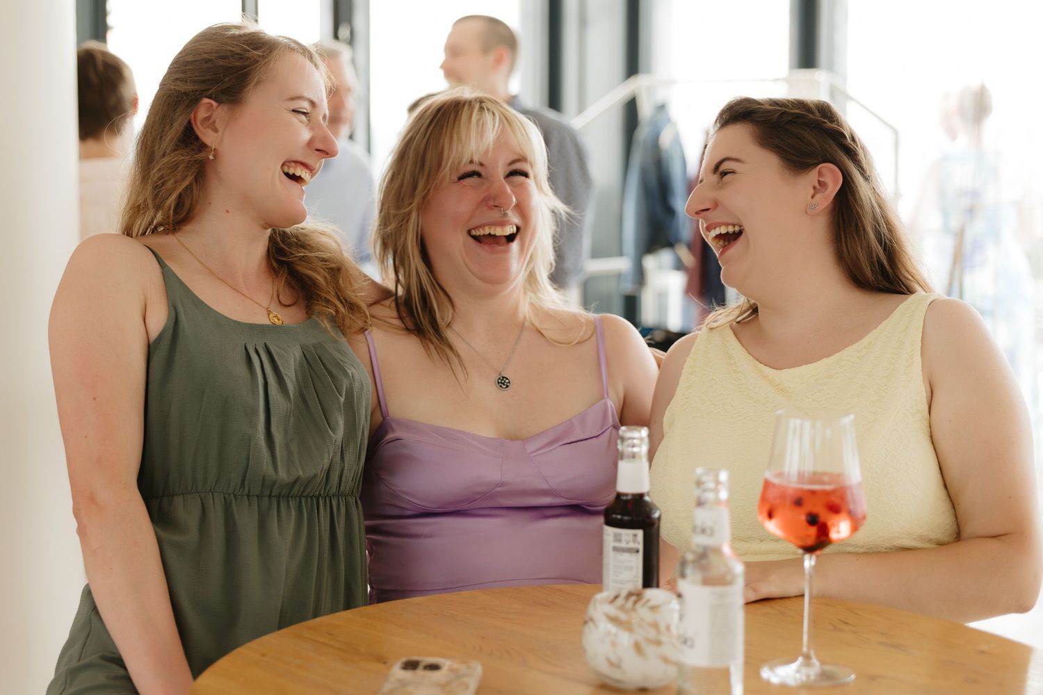 A group of women in summer dresses laugh and smile together during a social gathering at a bright indoor venue.