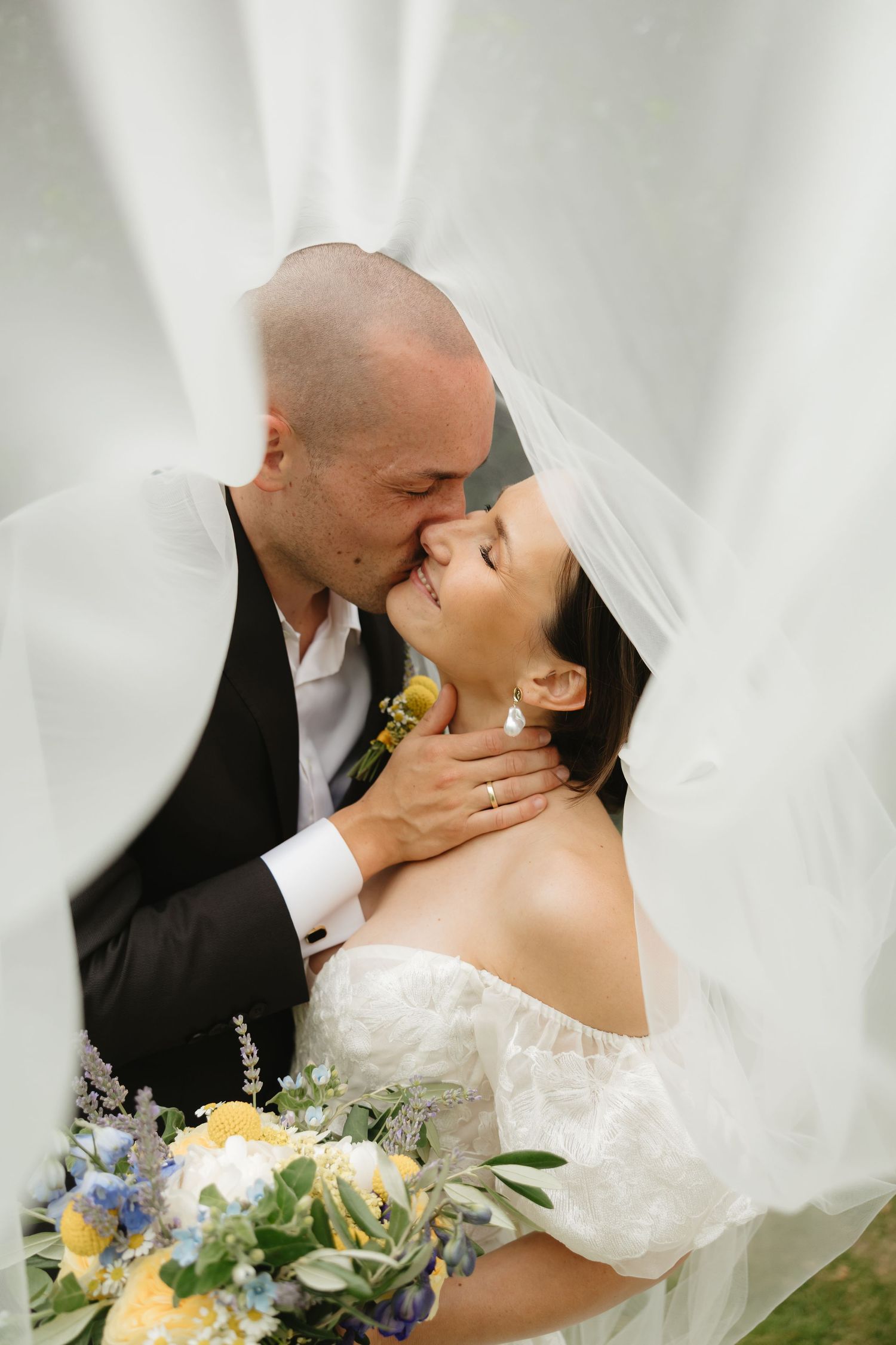 Bride and groom embrace in a passionate kiss while holding a colorful bouquet.