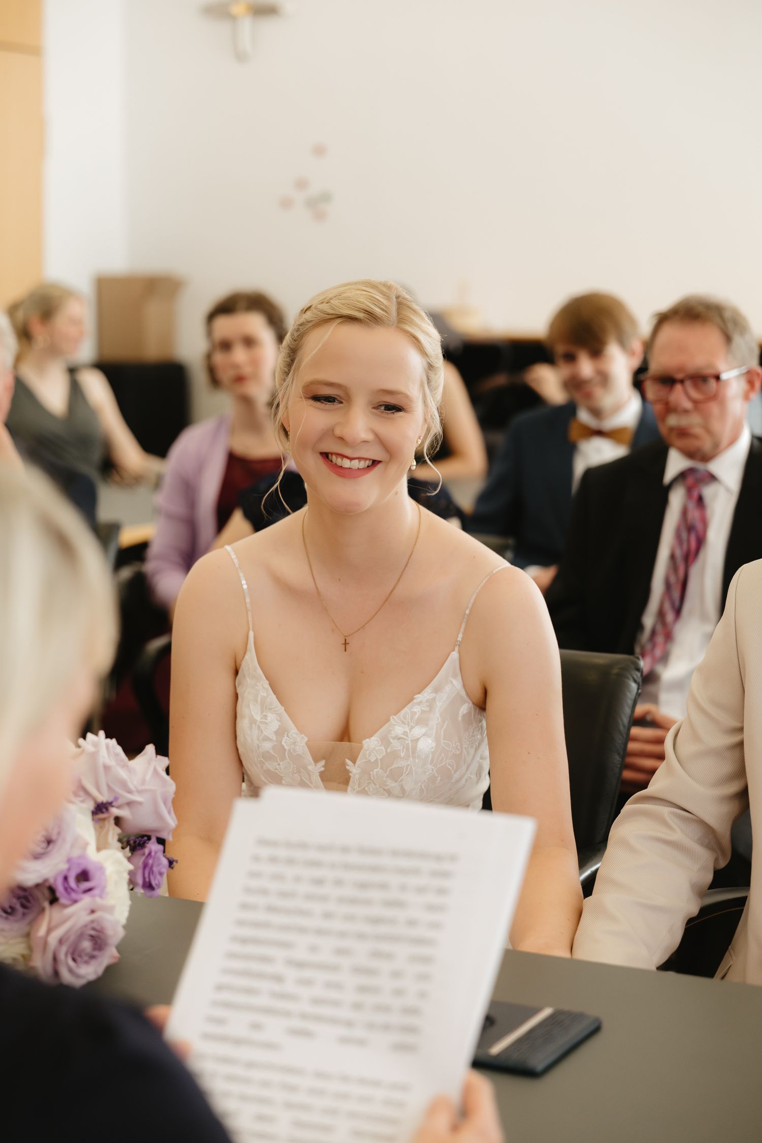 Bride in white lace dress smiles during wedding ceremony while document is held in foreground.