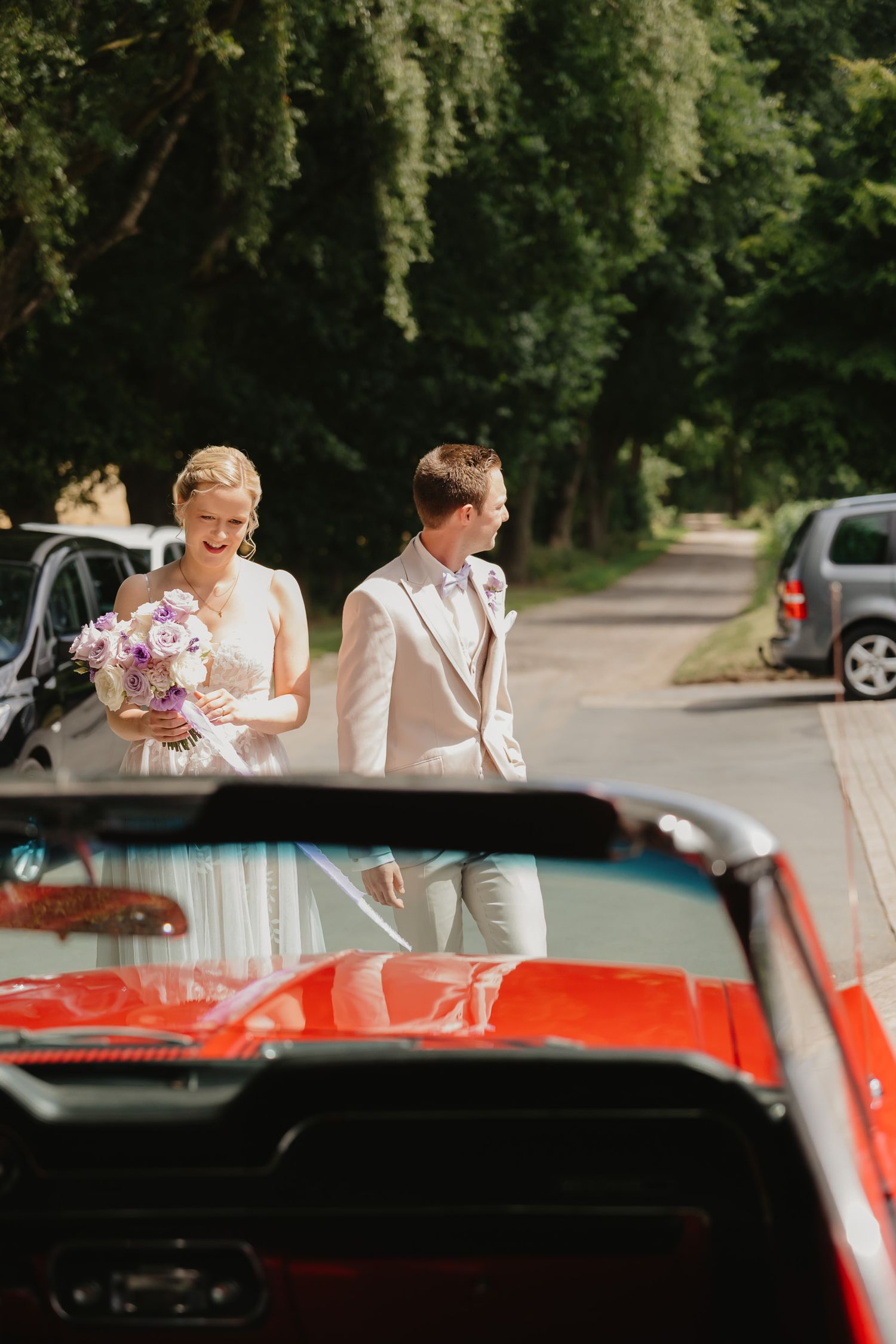 A red classic car with passengers is parked in a wooded area on a sunny day.