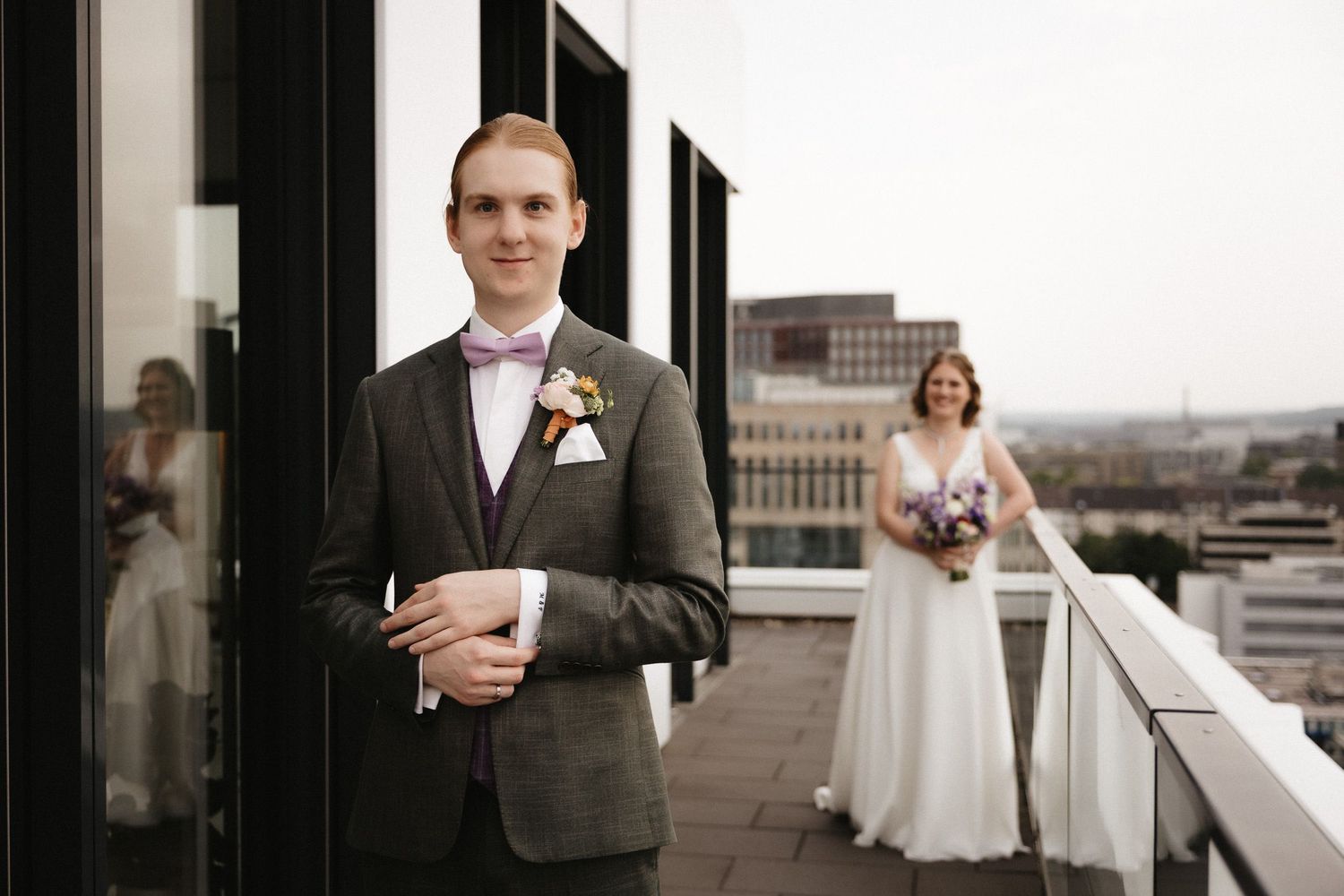 A wedding party stands on a rooftop balcony overlooking a city skyline on a cloudy day.