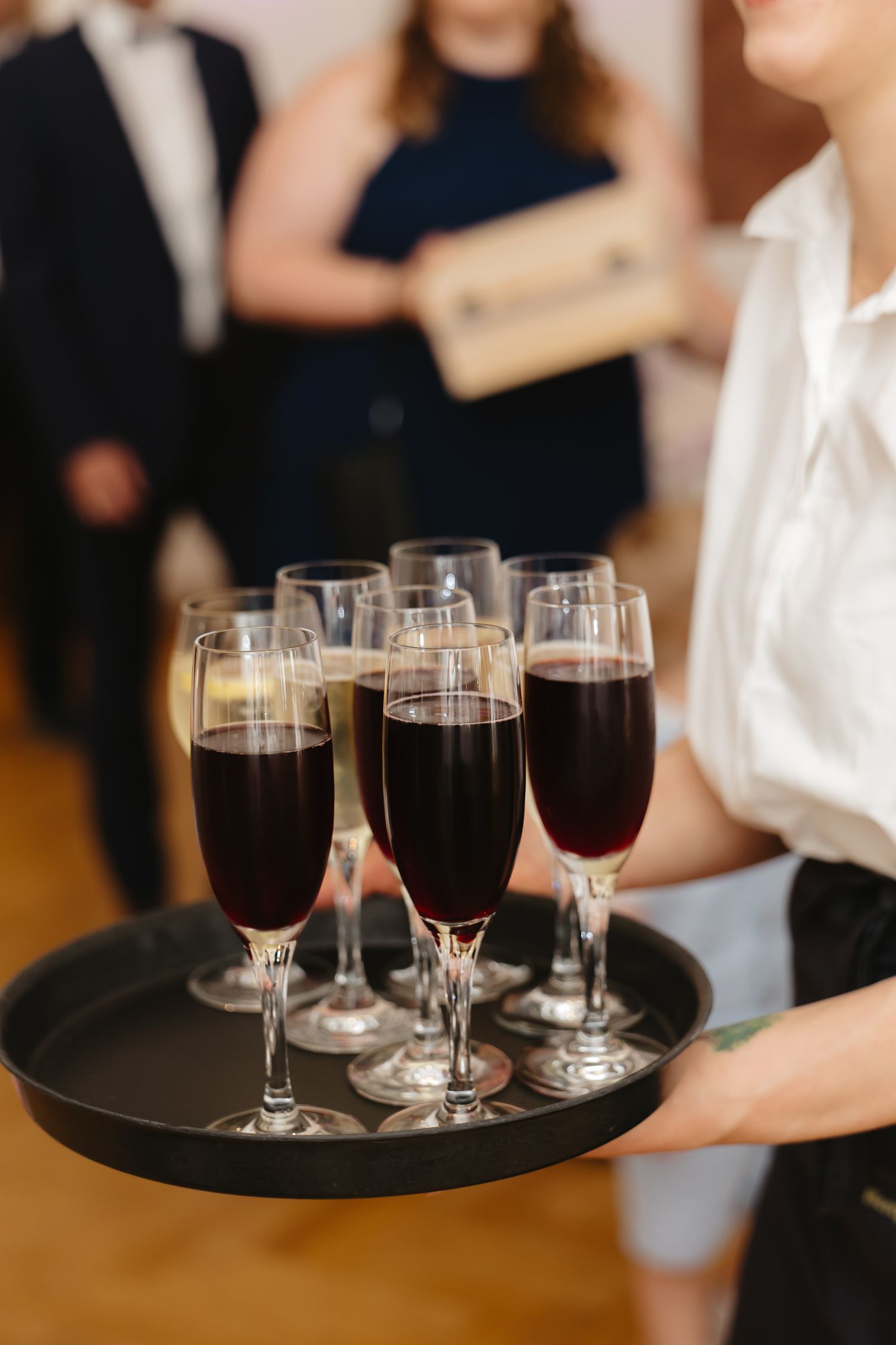 Server carries wine glasses filled with red wine on black serving trays at an elegant event.
