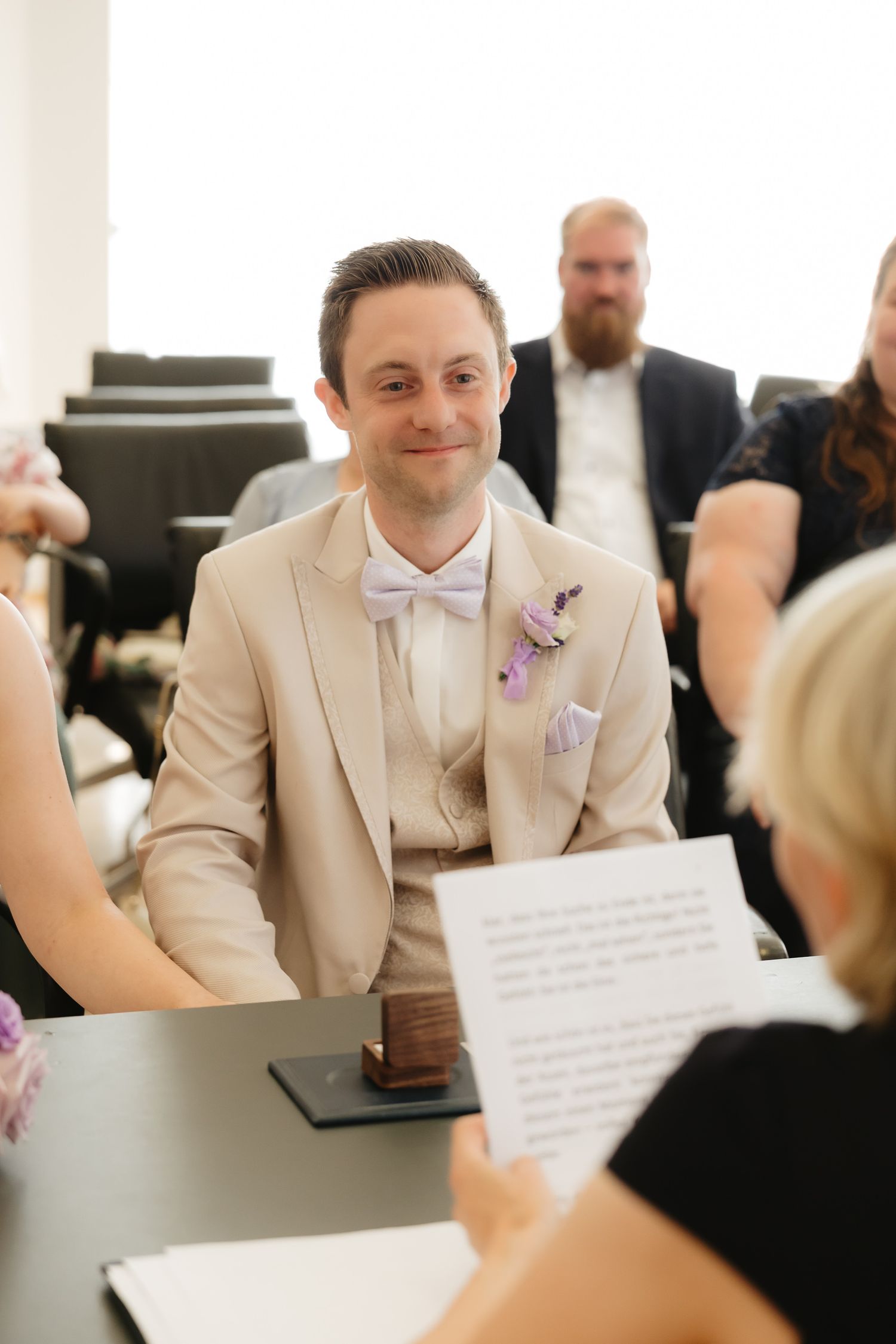 A person in a beige suit and white bowtie smiles during a formal event.