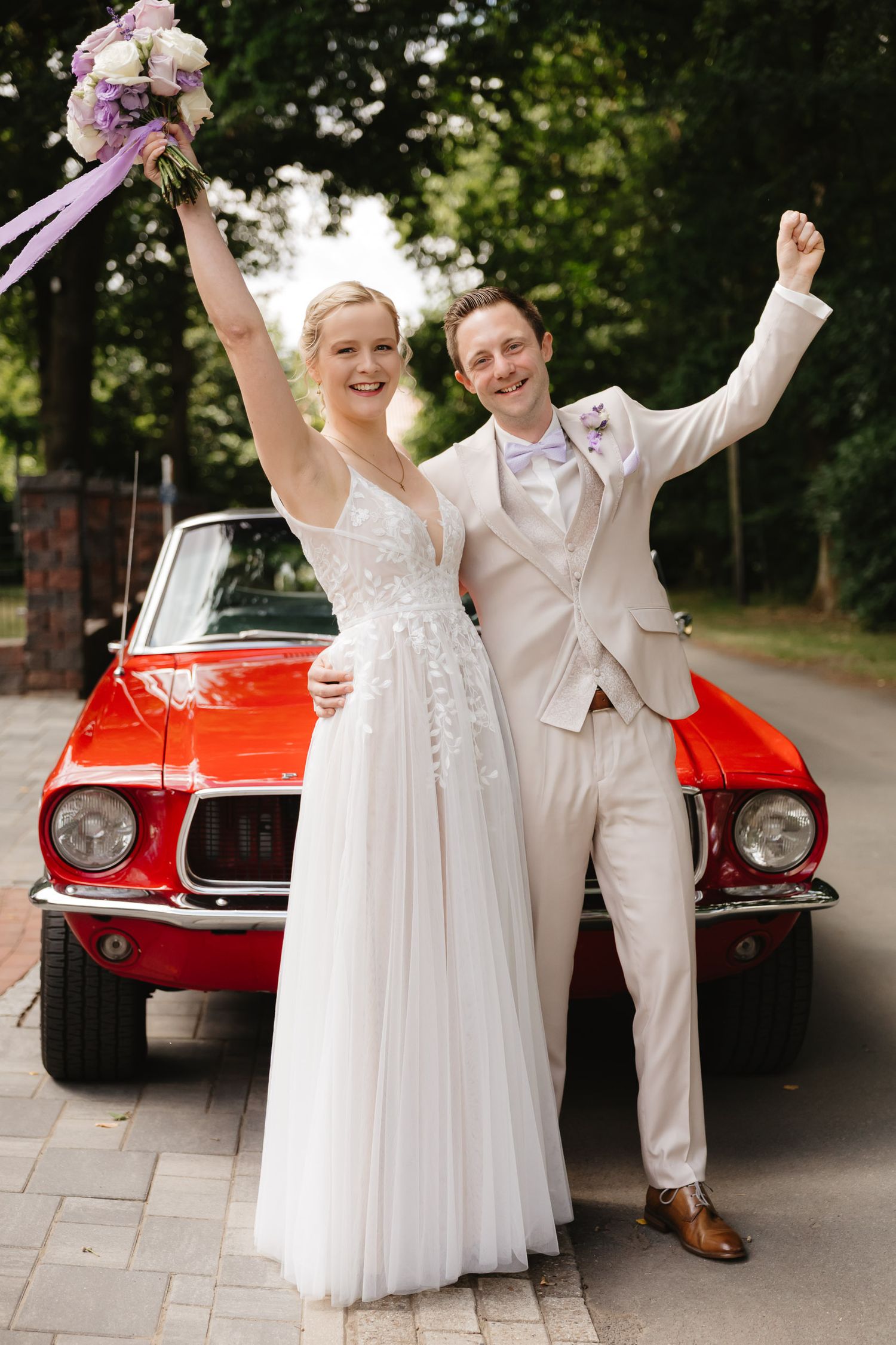 A couple in wedding attire poses joyfully in front of a vintage red car during their outdoor celebration.