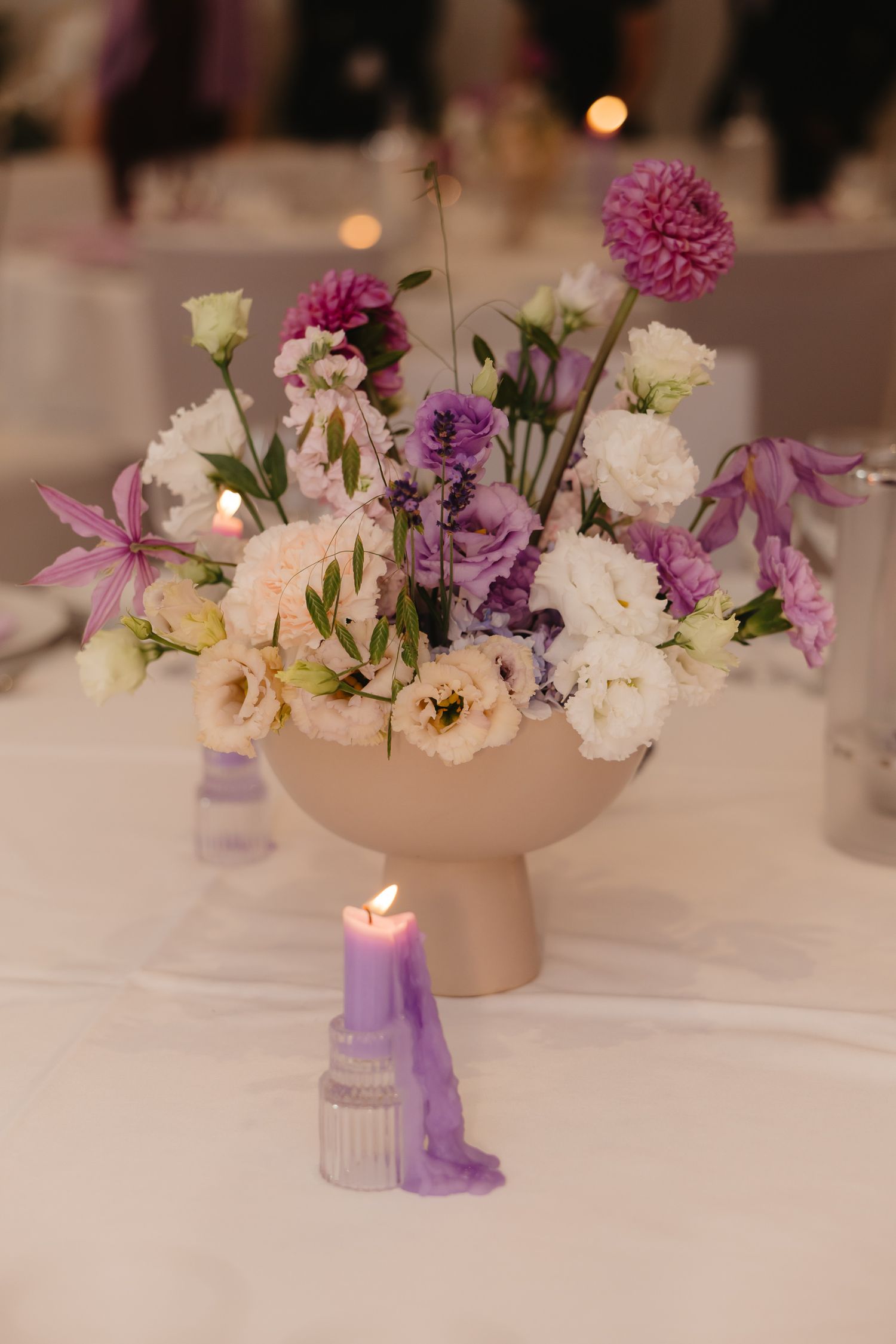 A delicate floral centerpiece with pink and white blooms in a cream pedestal vase, accompanied by a purple candle.