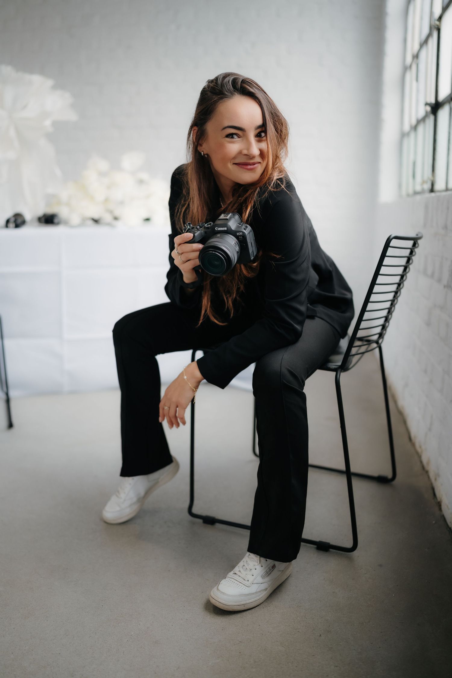 Professional photographer in black attire holding camera while sitting on chair in studio.