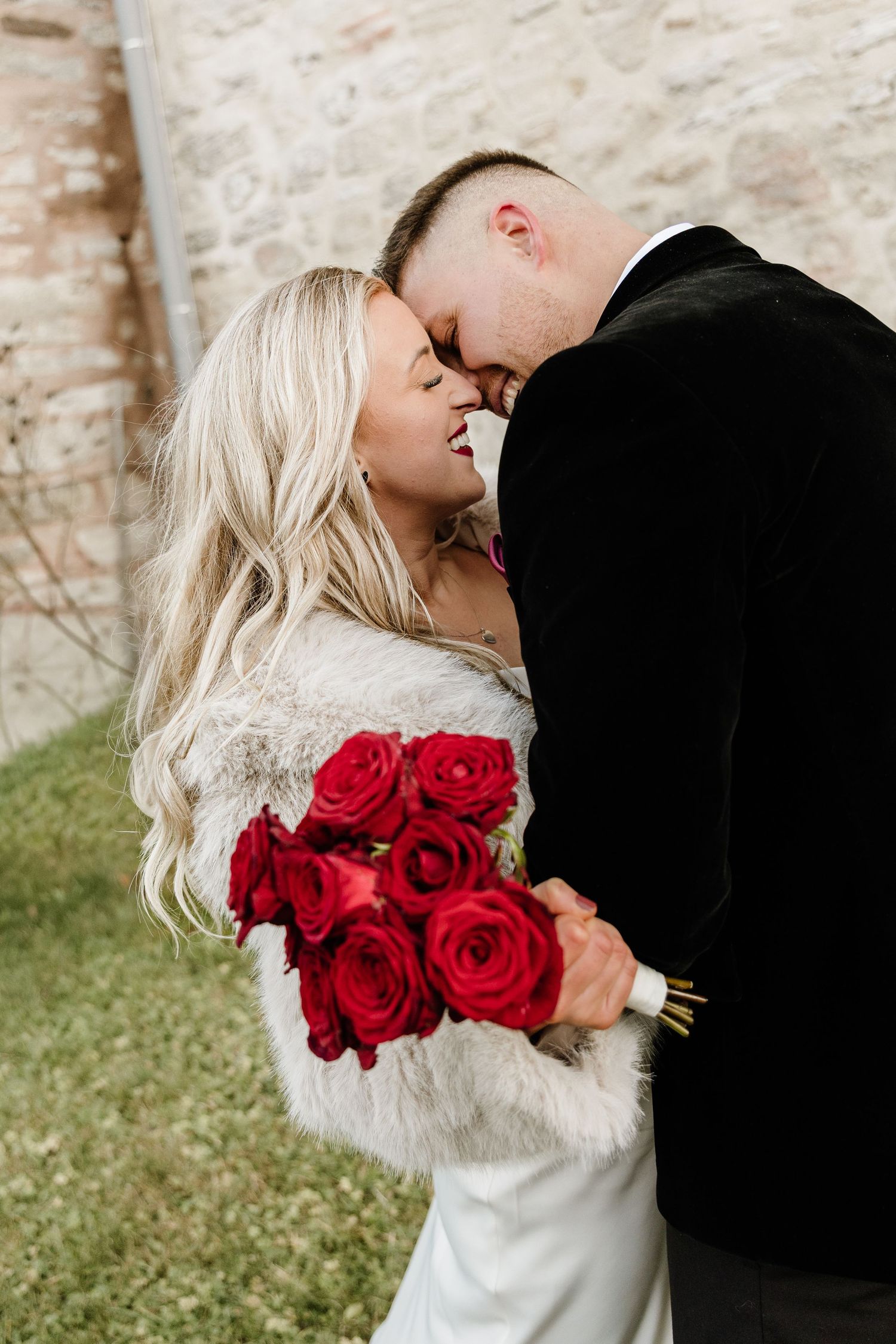 Couple shares intimate moment with red roses against stone wall backdrop.