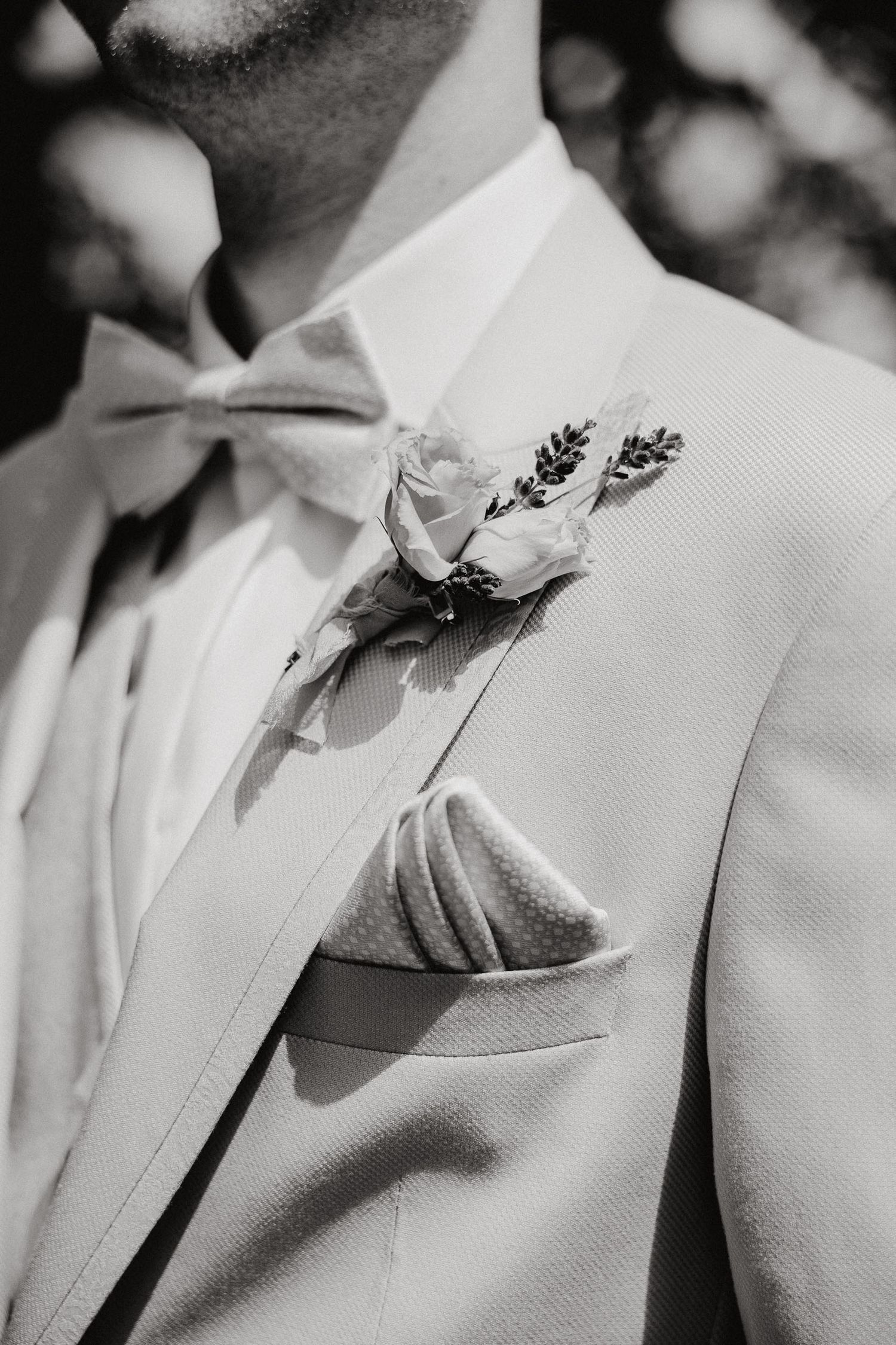 Close-up black and white photo of a light colored suit jacket with rose boutonniere and bow tie.