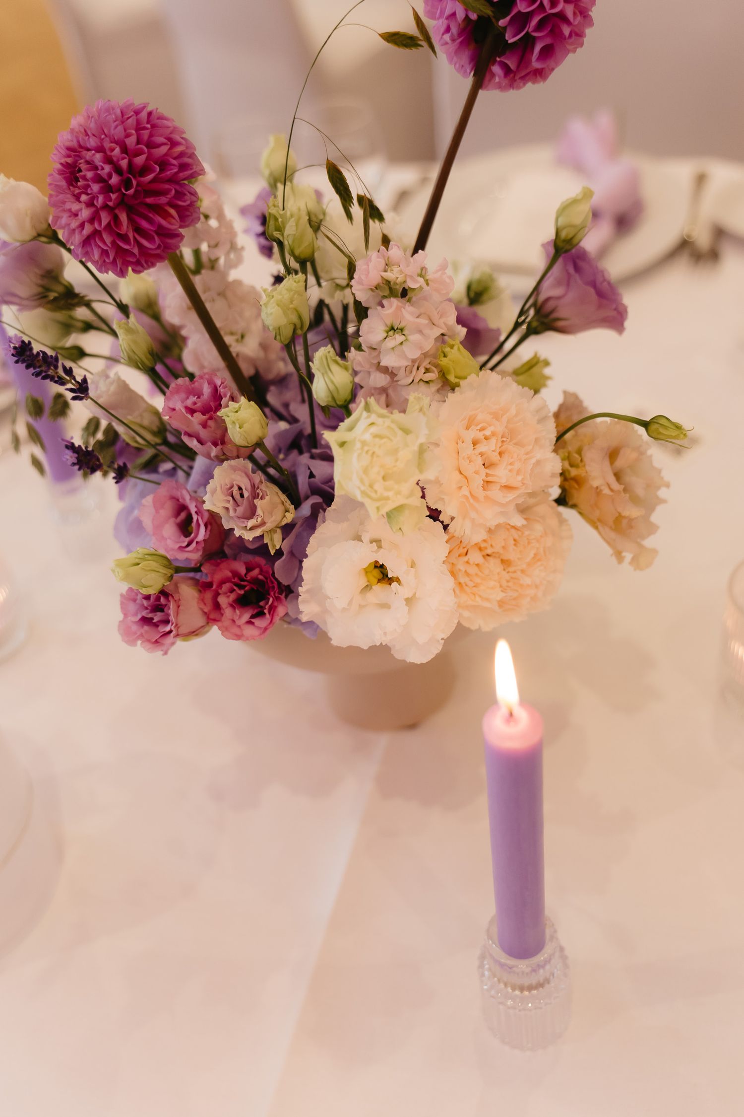 Romantic floral arrangement with purple and peach flowers beside a glowing lilac candle on a white tablecloth.
