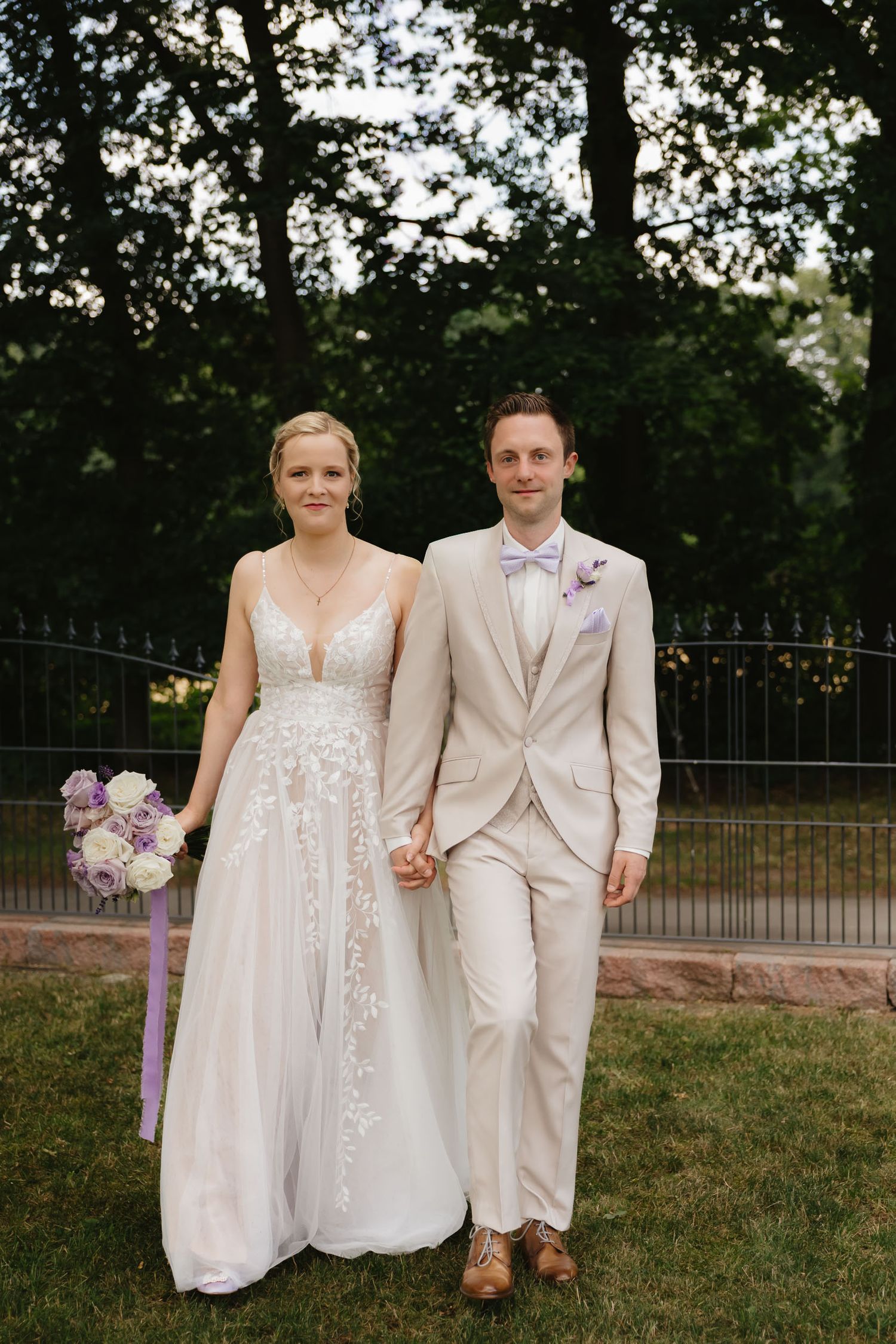 A couple in elegant wedding attire stands together on grass with trees and an iron fence in the background.