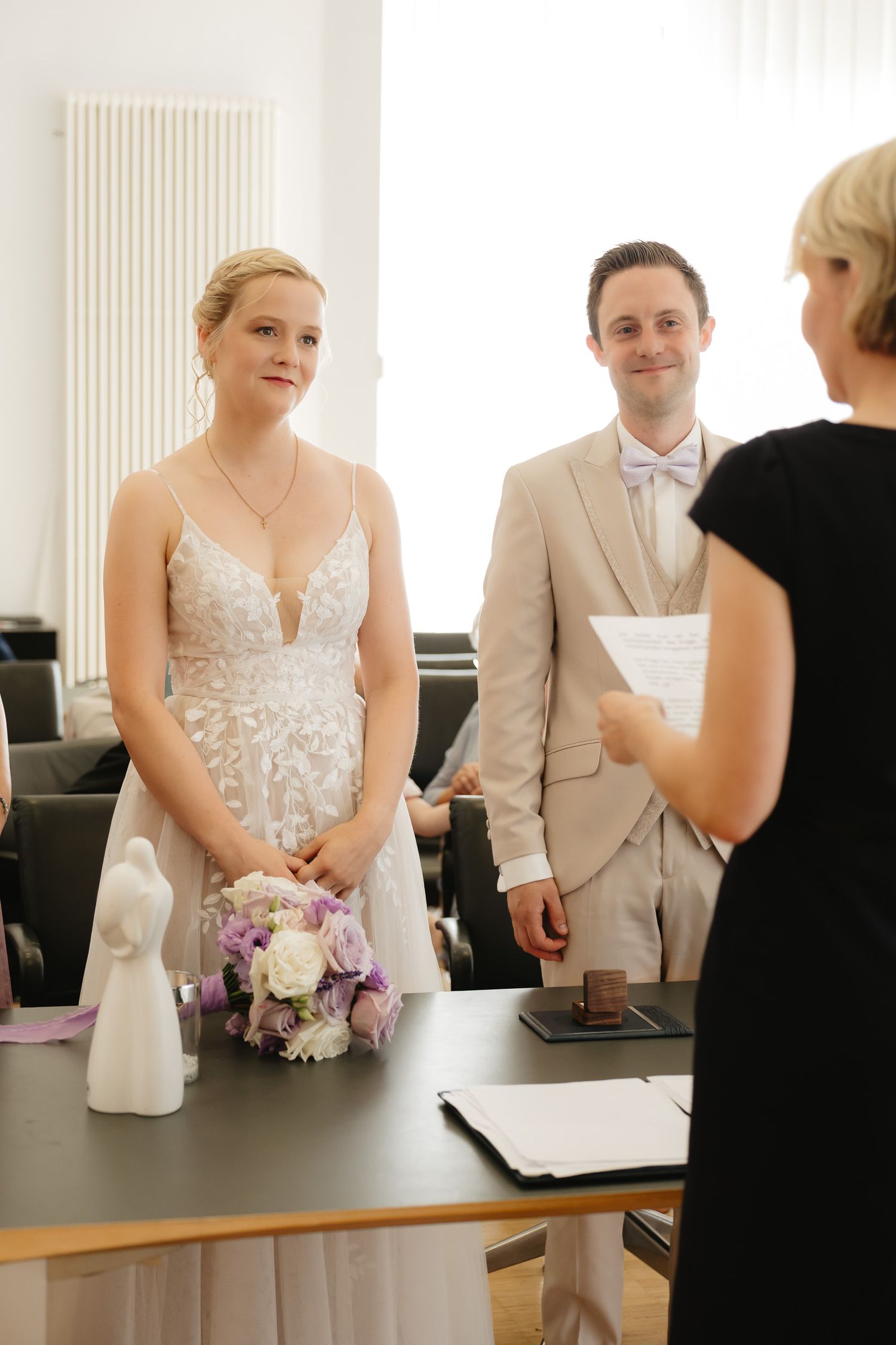 A couple stands at a registry desk during their civil wedding ceremony with a pink and white floral bouquet.