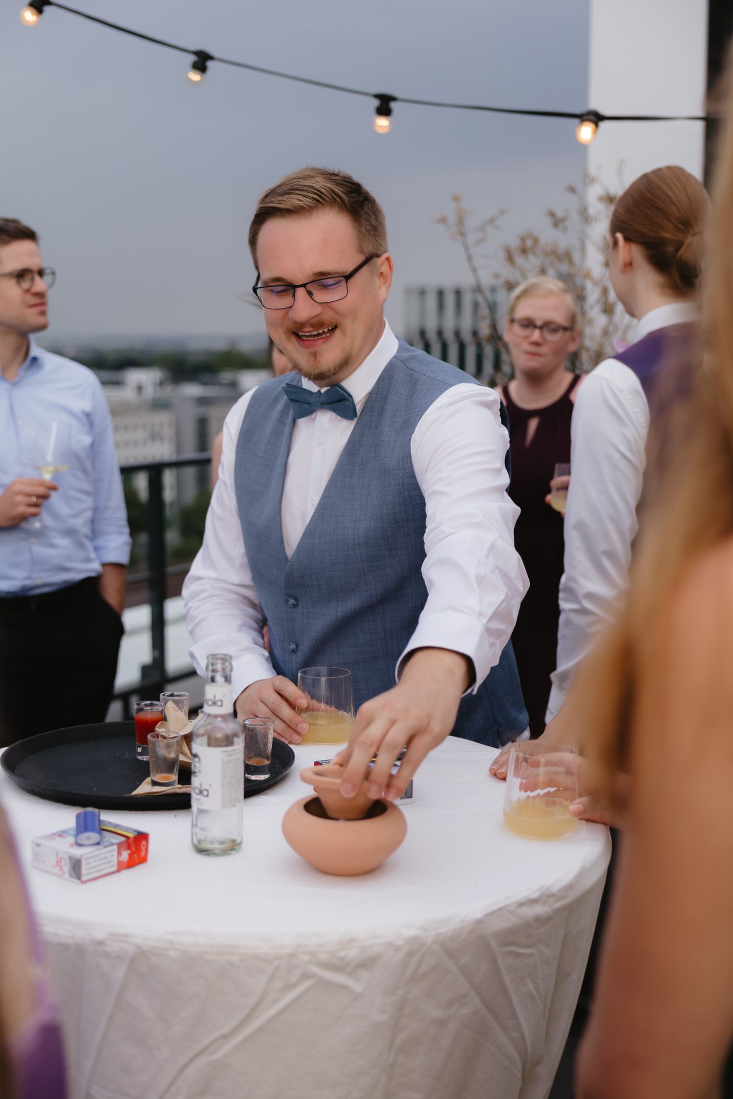 A person in a blue vest and bowtie serves drinks at an outdoor event with string lights overhead.