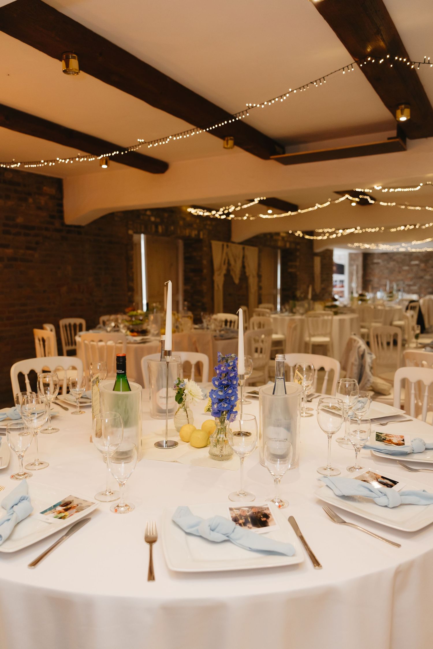 Elegant wedding reception tables set with white linens and string lights overhead in a rustic brick venue.