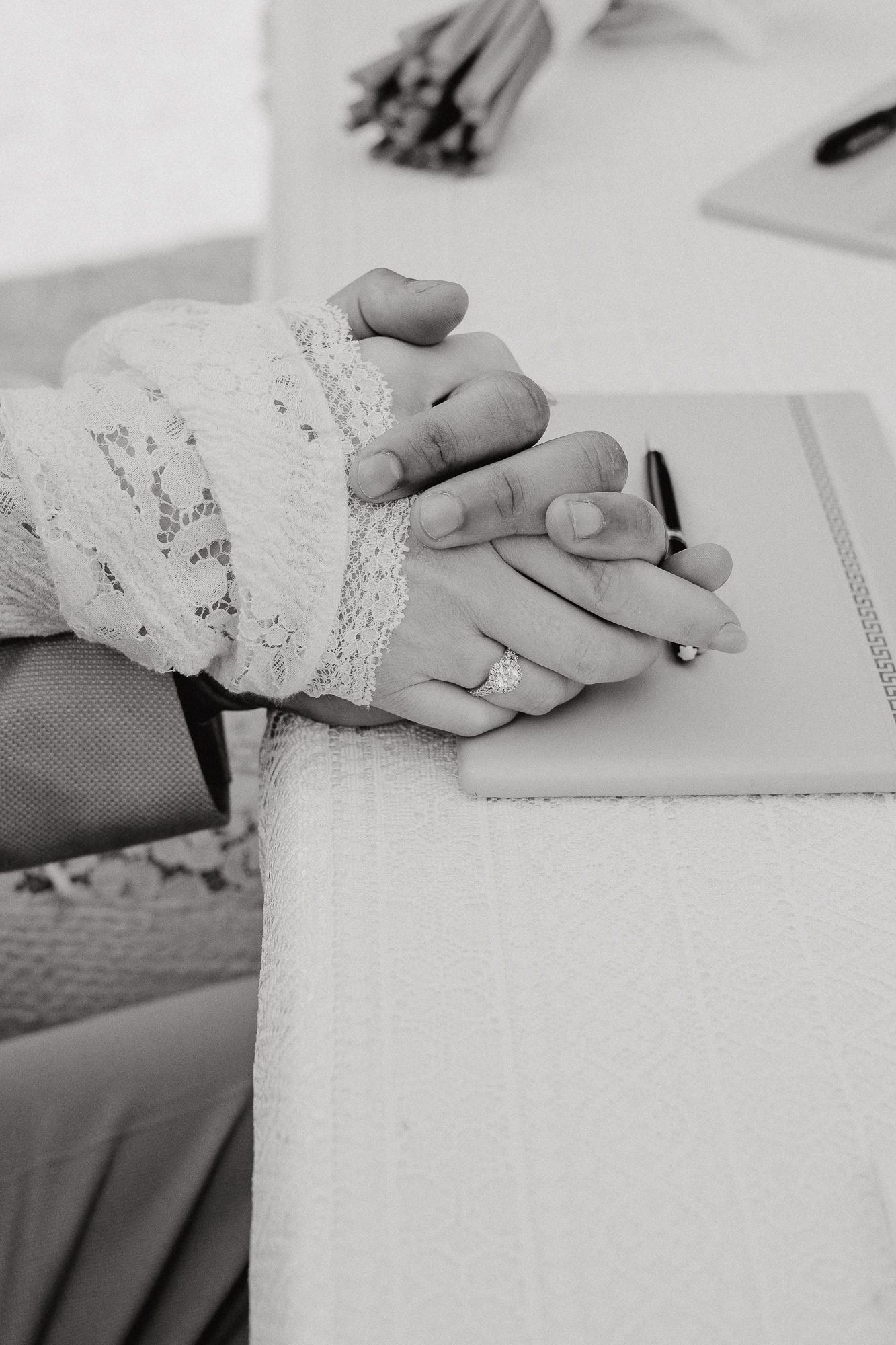 Black and white detail shot of lace-covered hands resting on a white surface during a wedding ceremony.