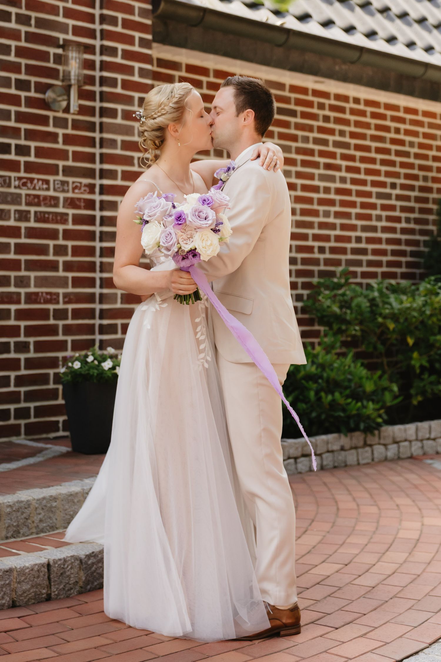 A romantic wedding couple shares a kiss while holding a purple and white bouquet against a brick building exterior.