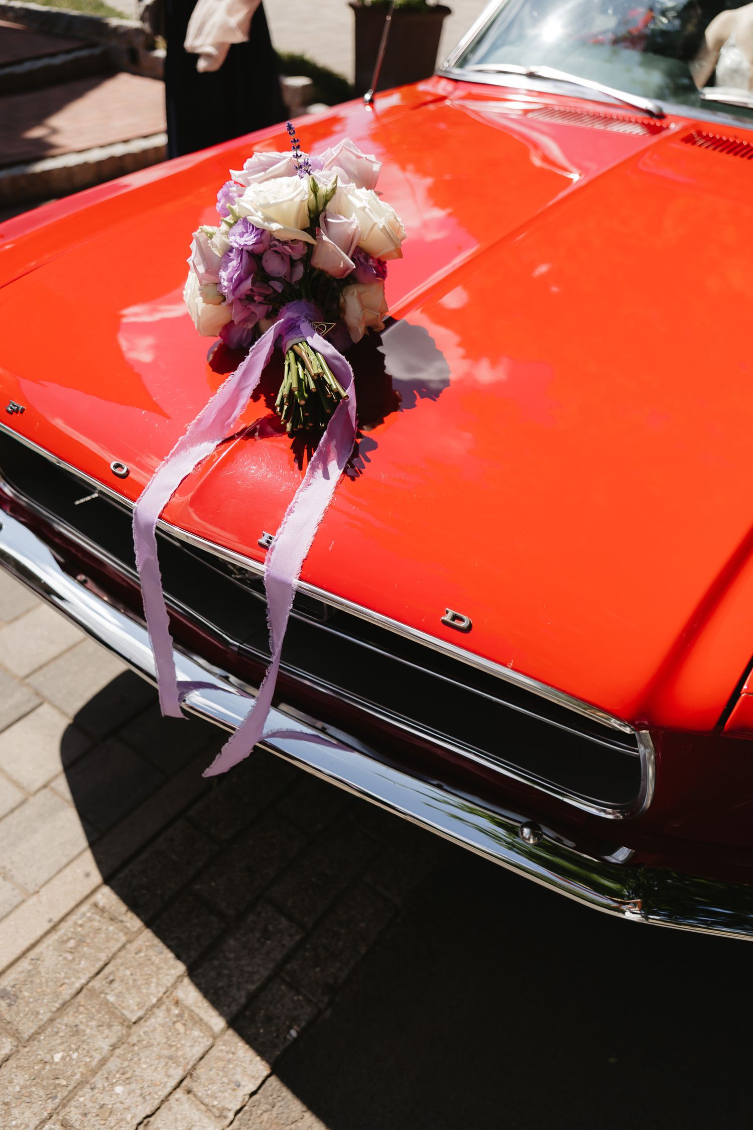 A wedding bouquet with purple and white flowers rests on the hood of a bright red classic car.