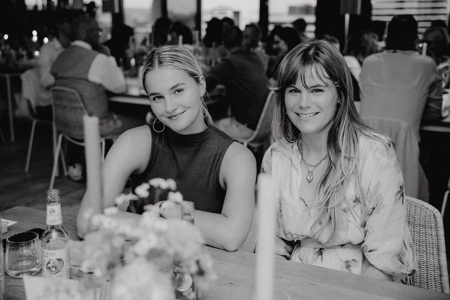 Two women sit together at a dining table in a black and white photo with floral centerpiece decorations.