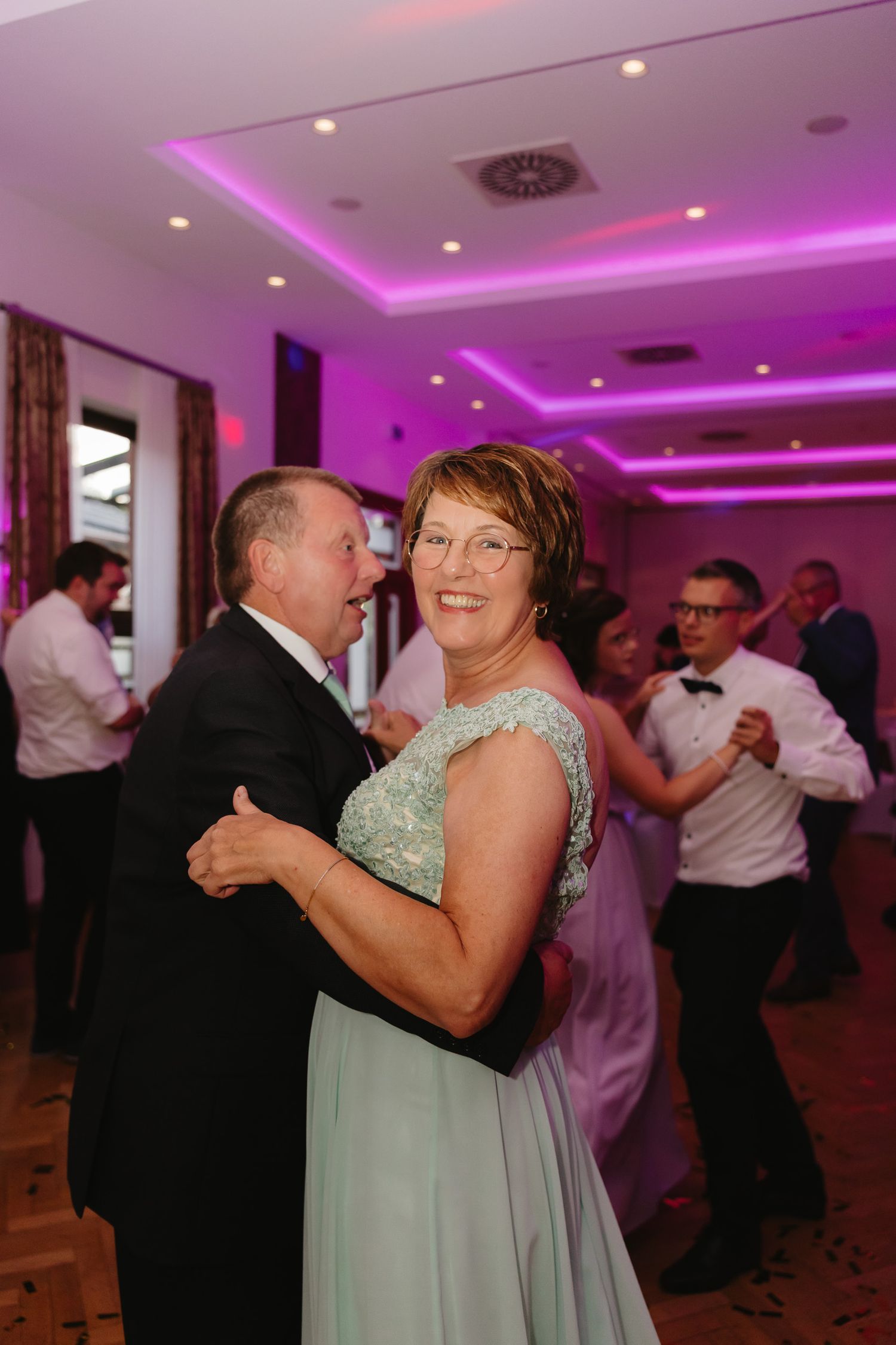 Couples dance together at a wedding reception with purple mood lighting illuminating the venue.