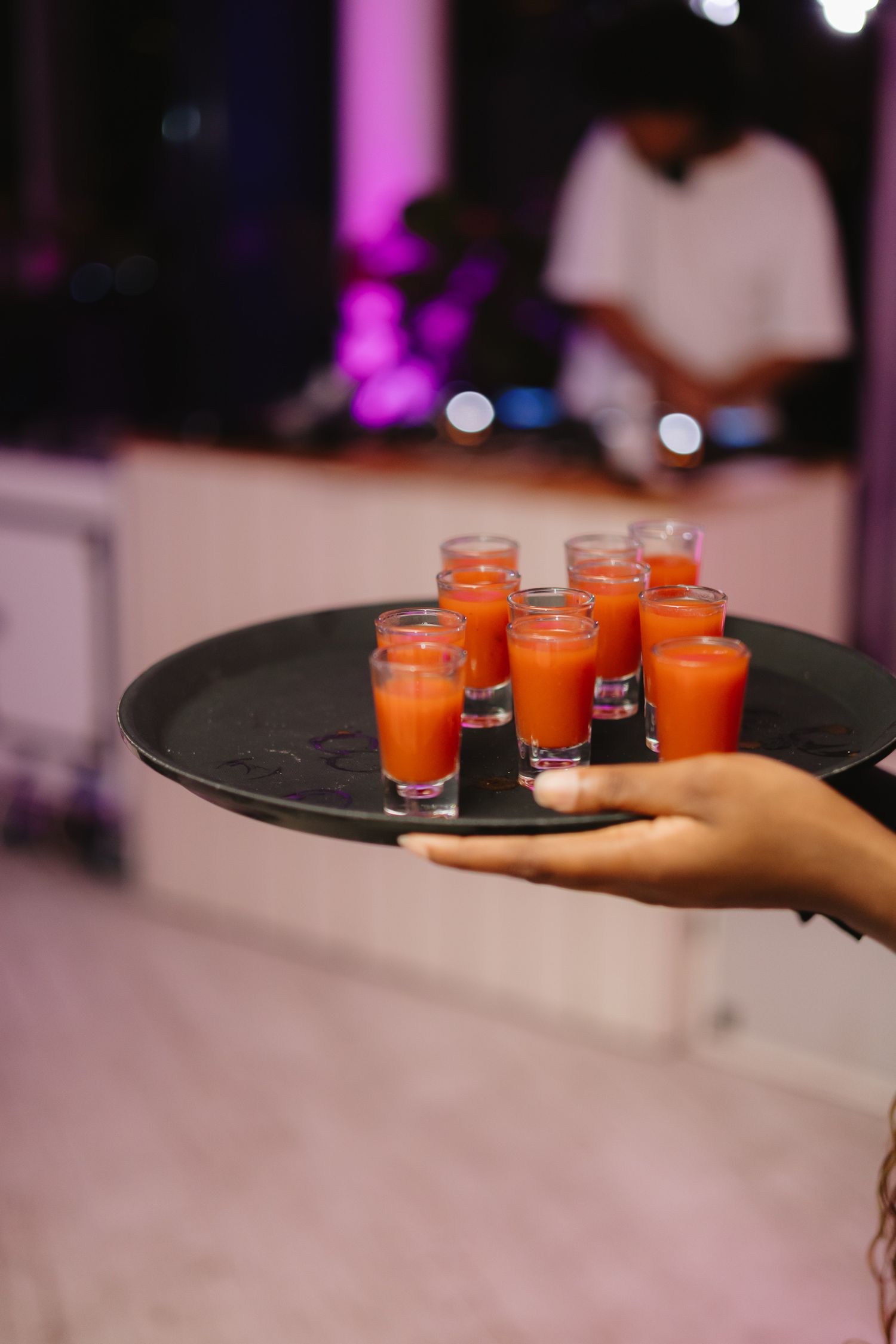 A hand holds a black serving tray with small glasses filled with bright orange cocktails in a dimly lit venue.