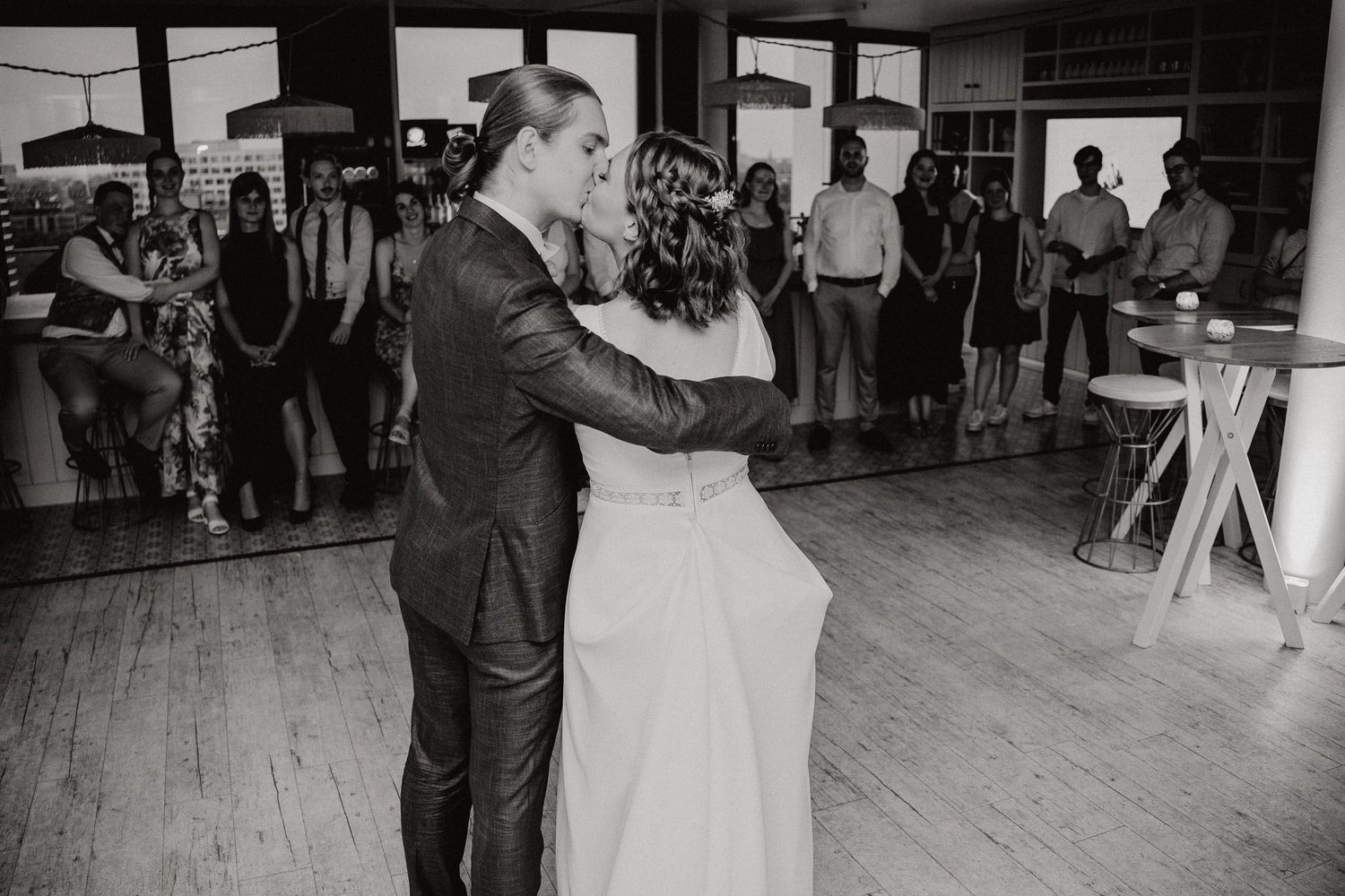 A couple shares their first dance at a rustic indoor wedding reception while guests watch from the perimeter.