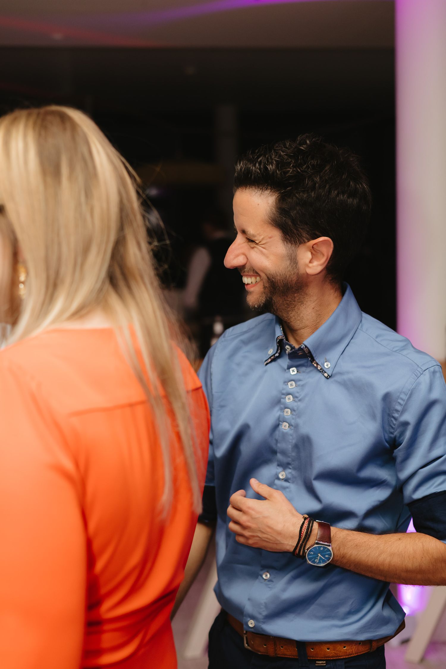 A couple in orange and blue attire share a candid moment during a conversation in a dimly lit setting.