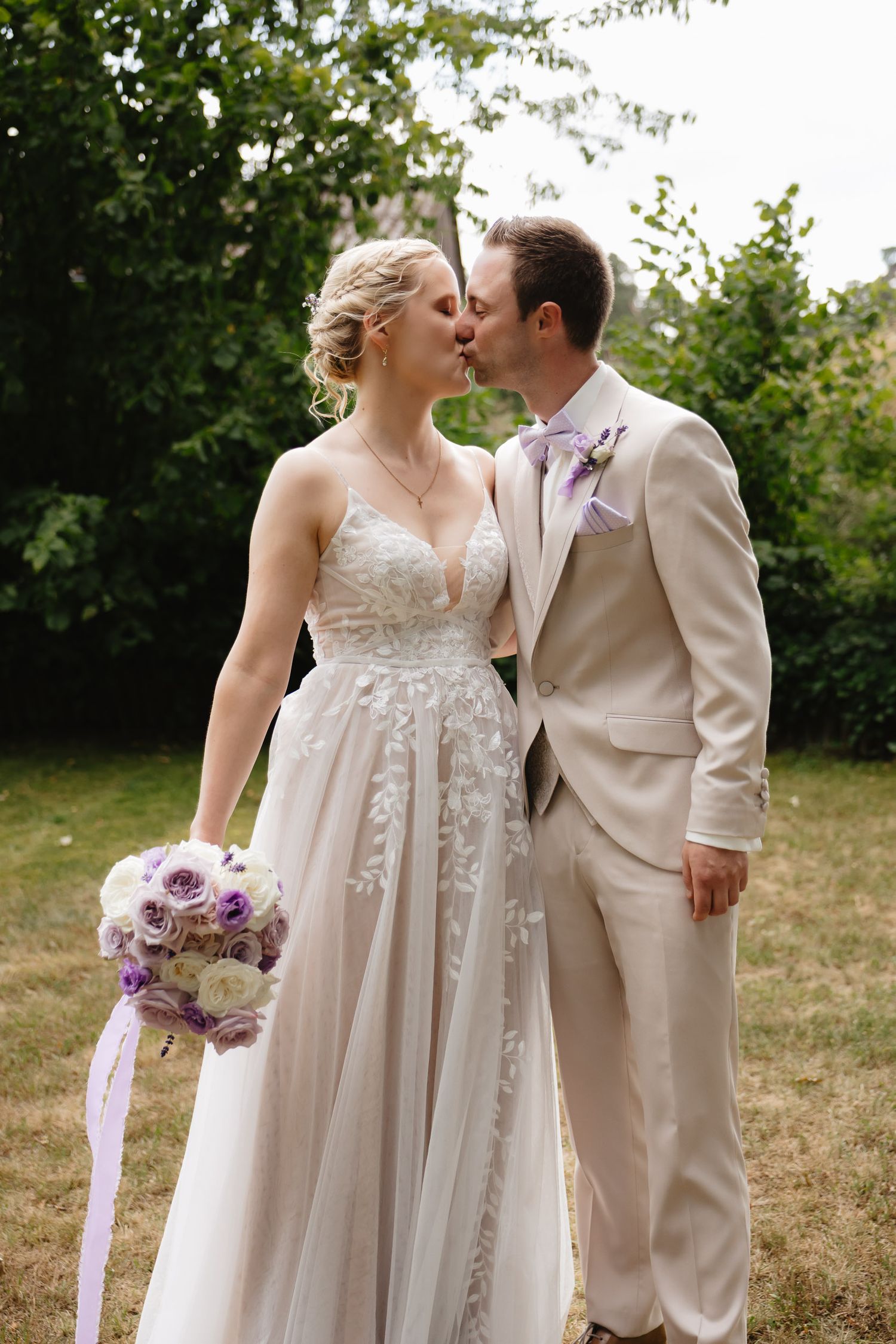 A romantic outdoor wedding moment with a couple in beige attire and a purple bouquet in a garden setting.