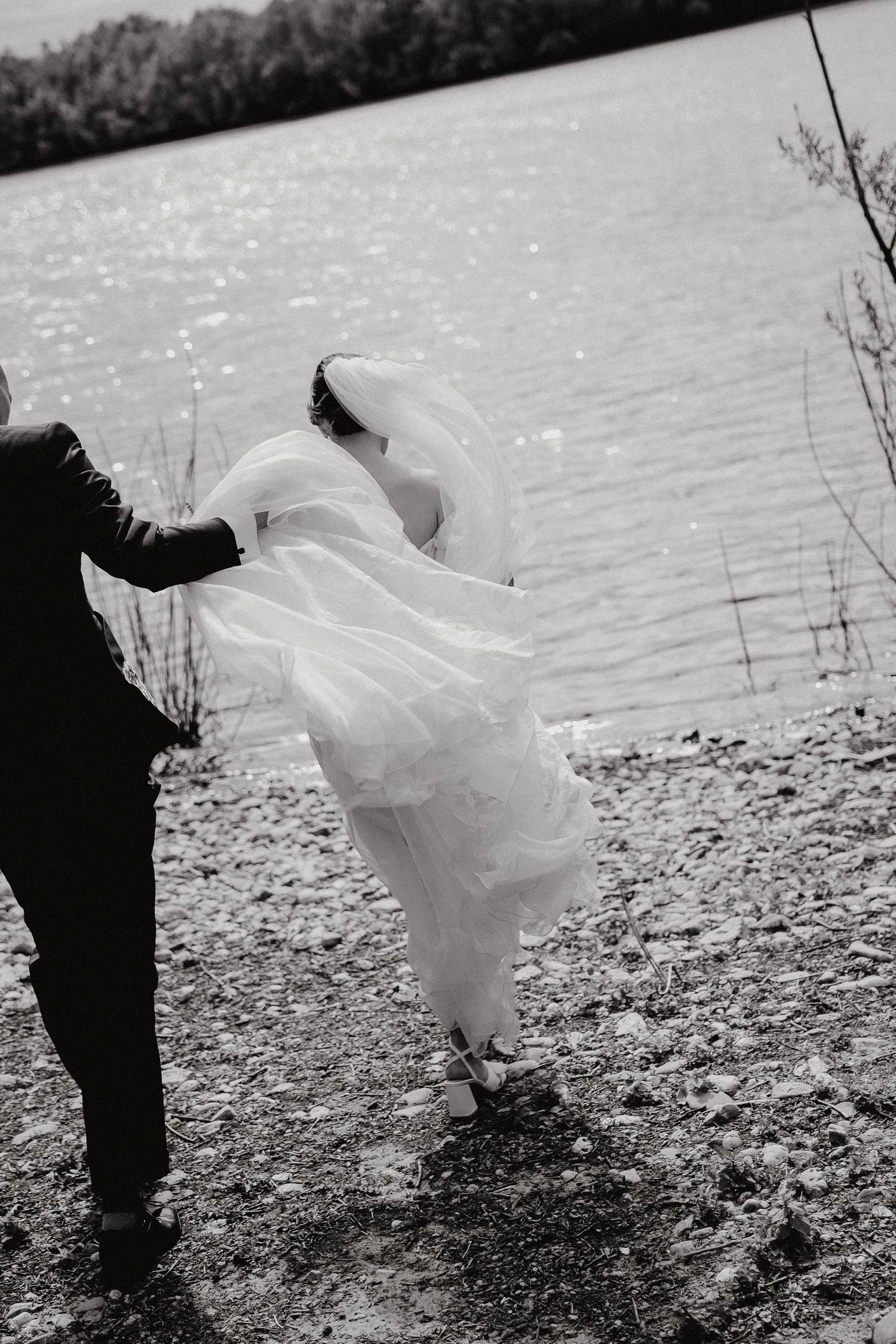 Silhouetted couple in wedding attire pose by a sparkling lake.