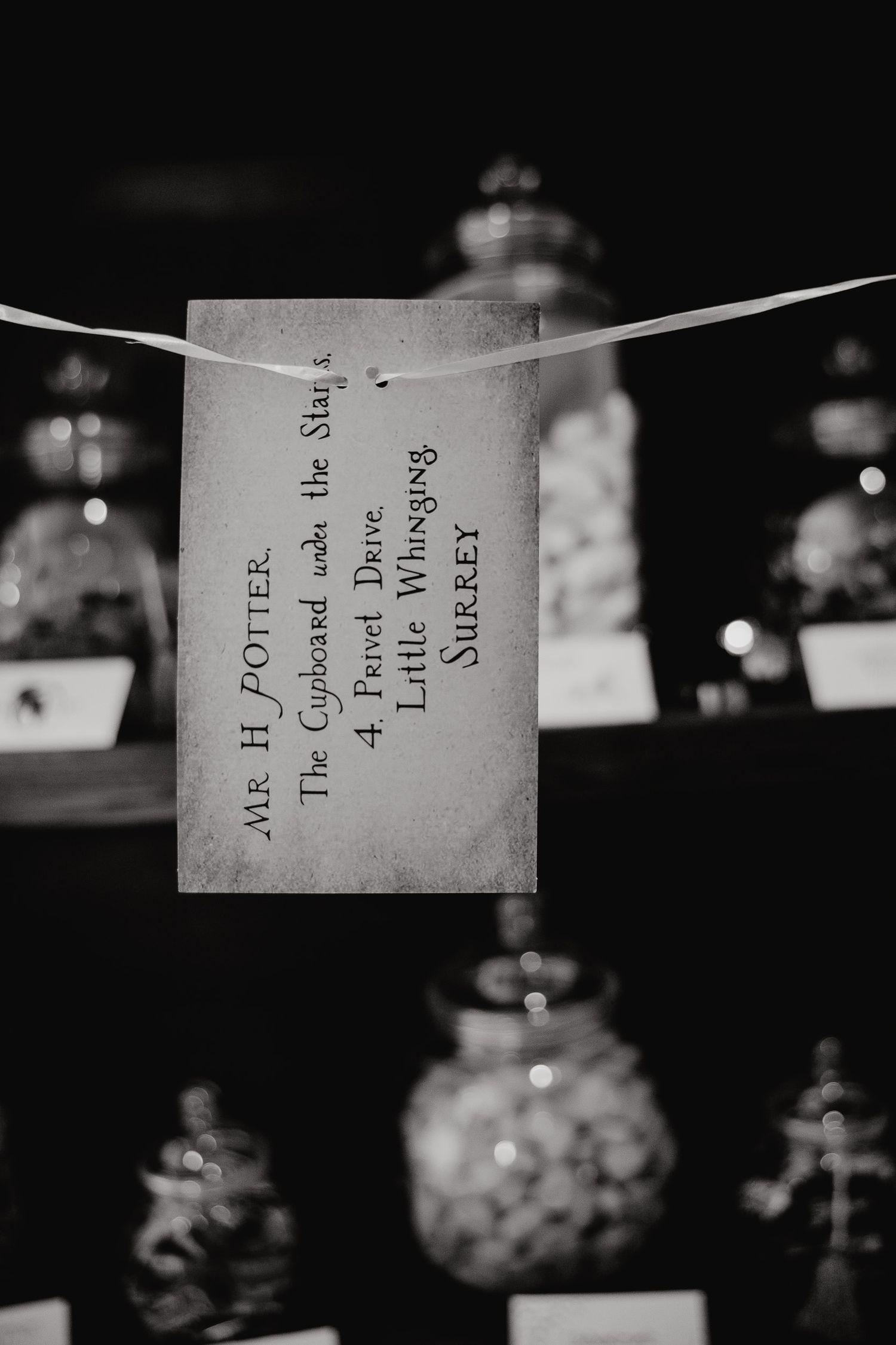 Black and white photo of a handwritten note hanging on a string with glass jars visible in the background.