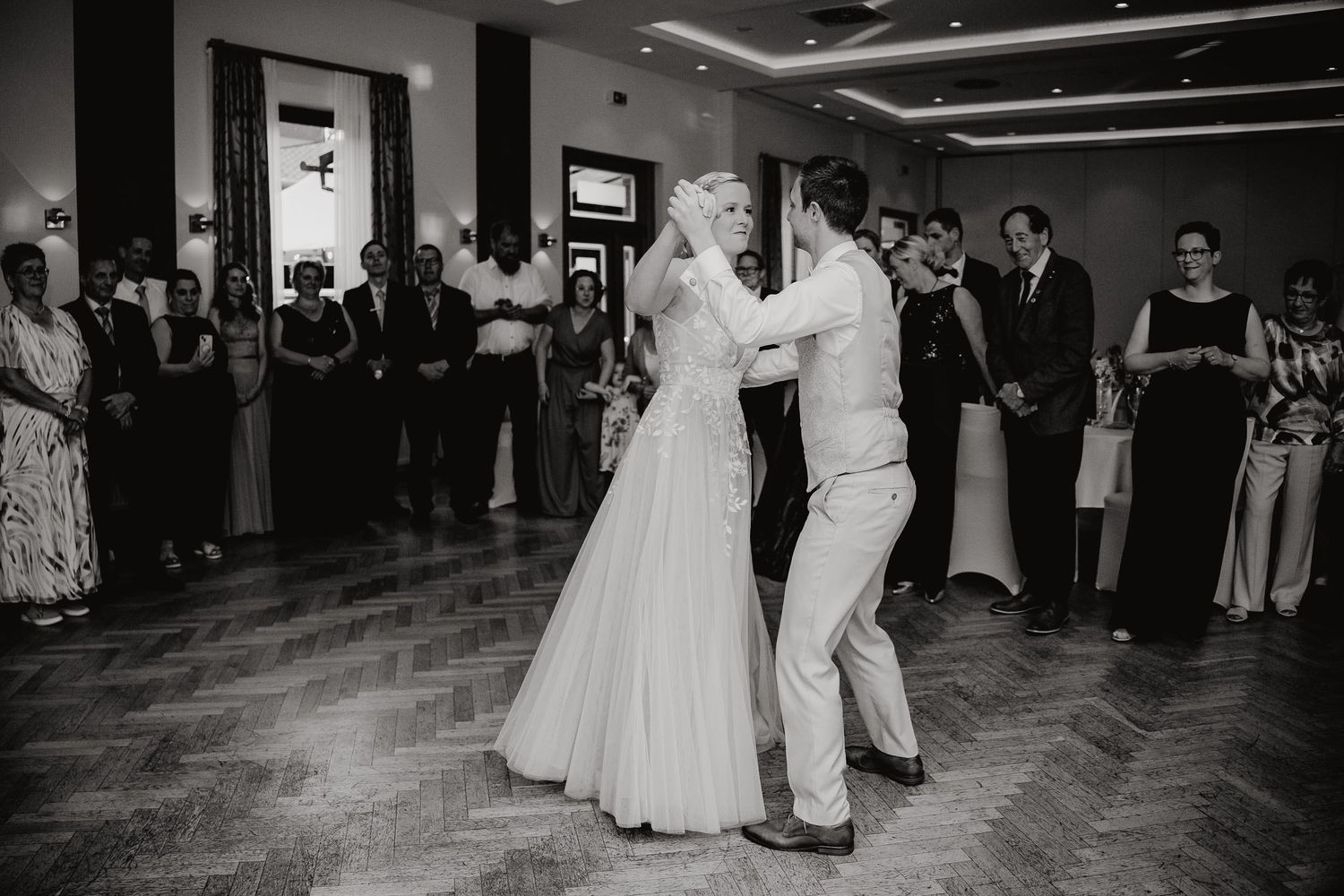 A couple shares their first dance at a wedding reception on a wooden dance floor while guests watch from the sidelines.