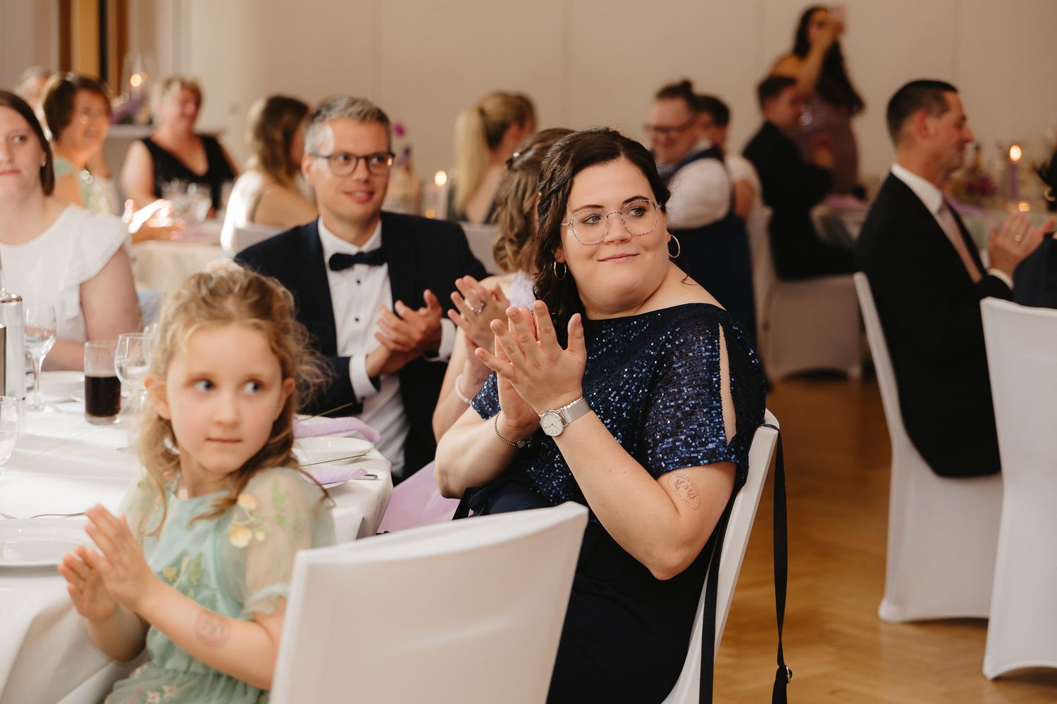 Wedding guests clap and celebrate during a reception in an elegant indoor venue with warm lighting.