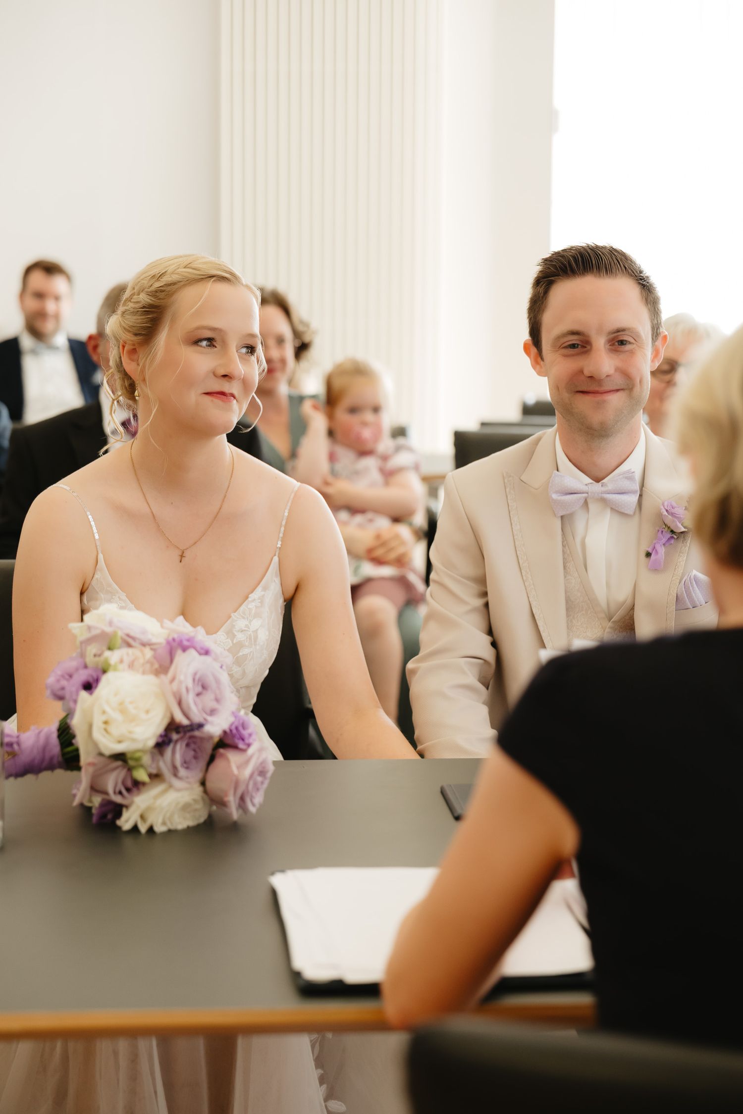 Wedding party members in beige suits and dresses sit at a reception table with purple and white floral bouquets.