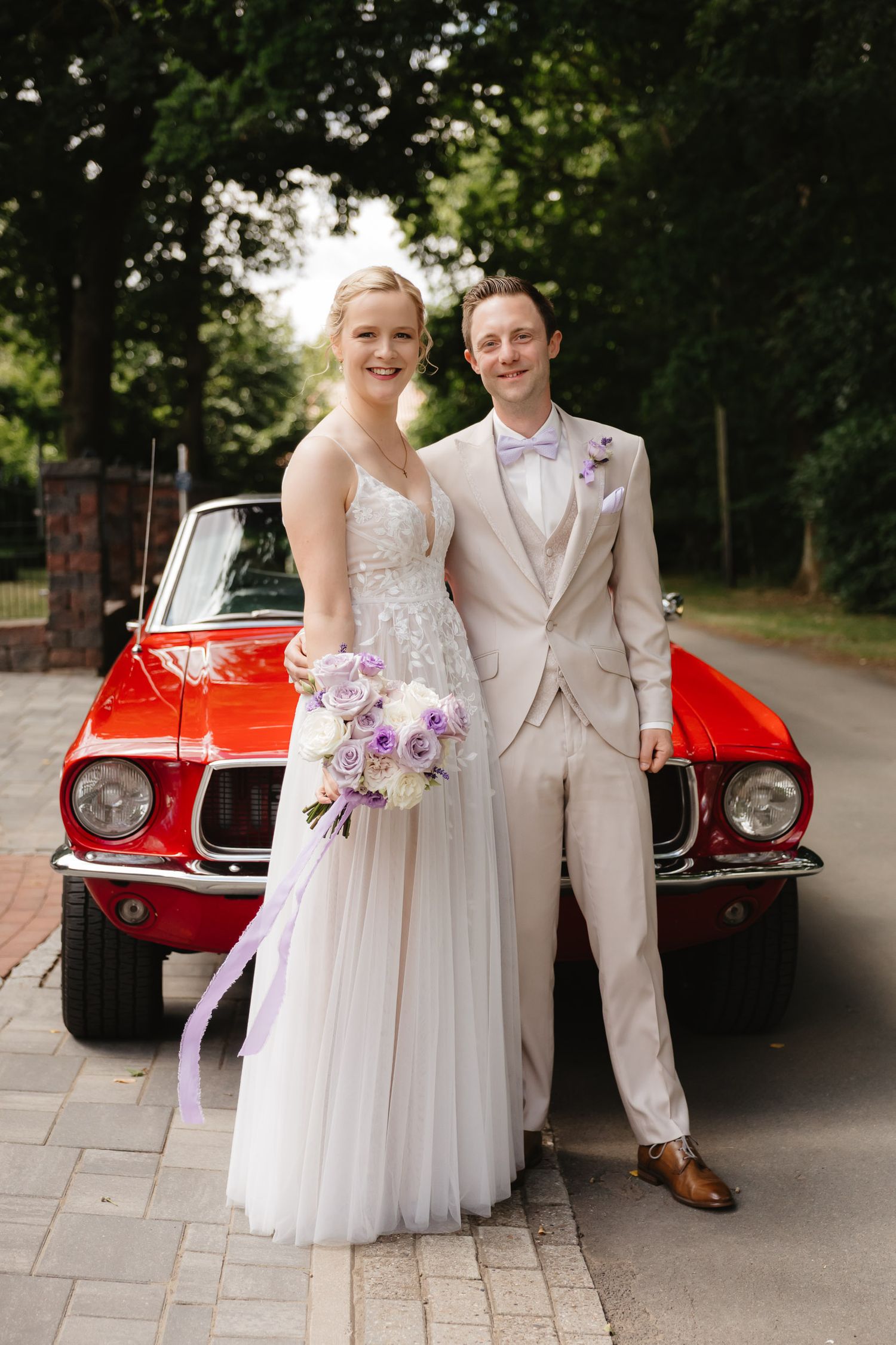 A couple poses in wedding attire in front of a classic red Mustang car on a tree-lined street.