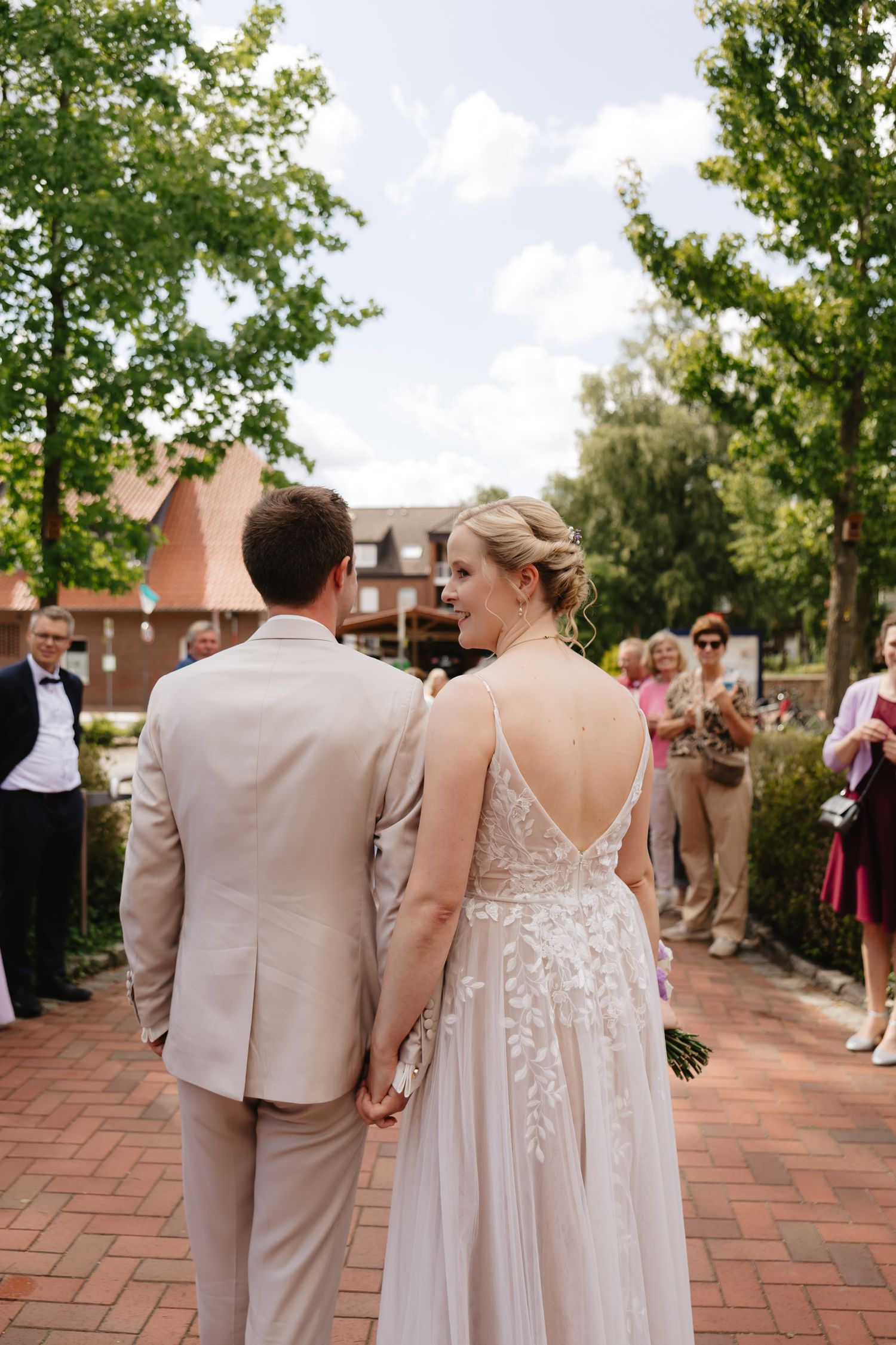 Wedding party members in blush pink dresses and tan suits walk along a brick pathway during an outdoor celebration.