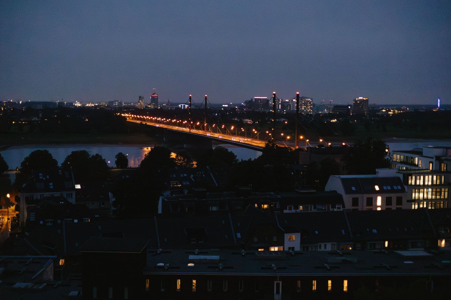 Illuminated bridge spans across river at dusk with city skyline and residential buildings in Minneapolis.