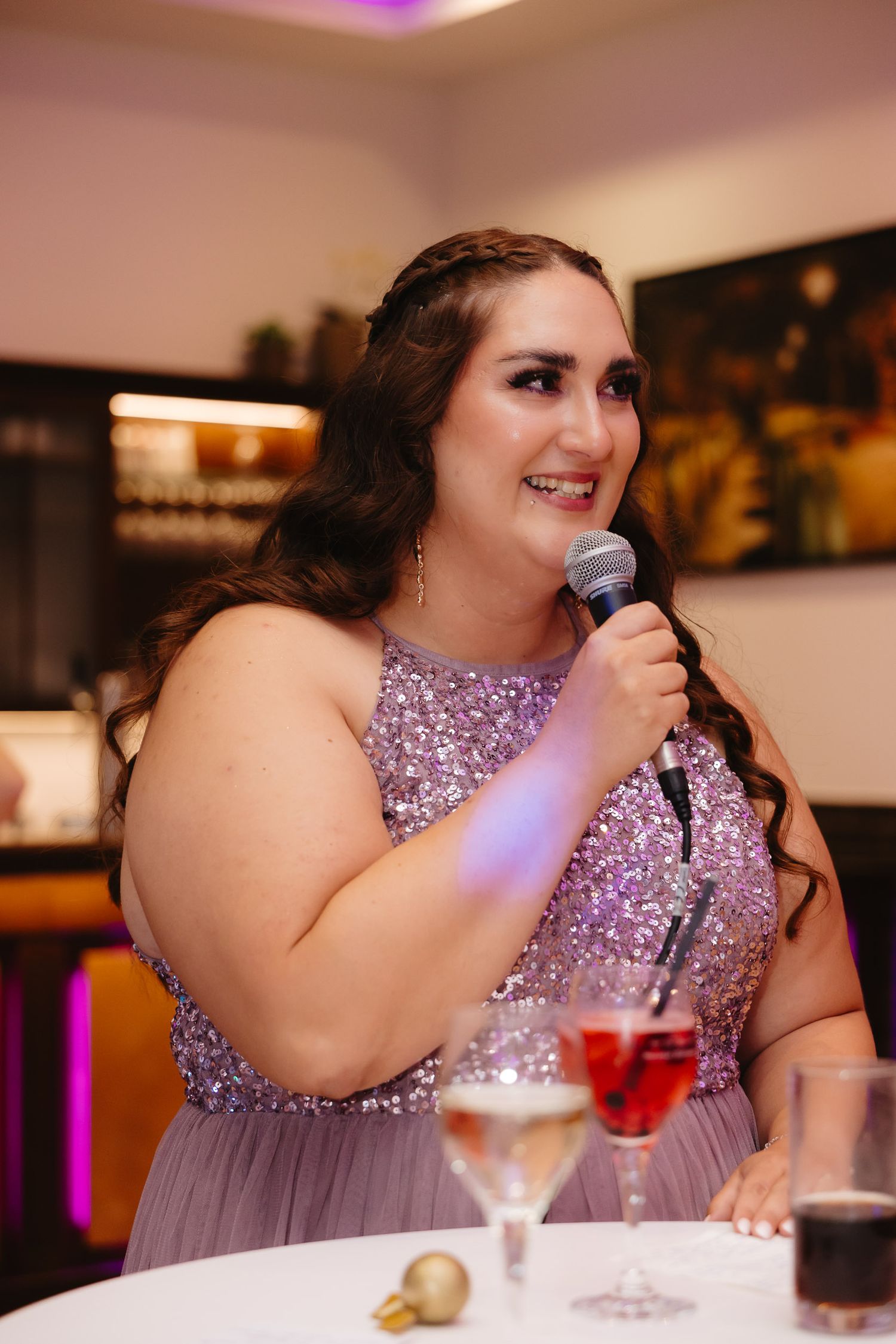 A person in a sparkly purple dress speaks into a microphone at a formal event with drinks on the table.