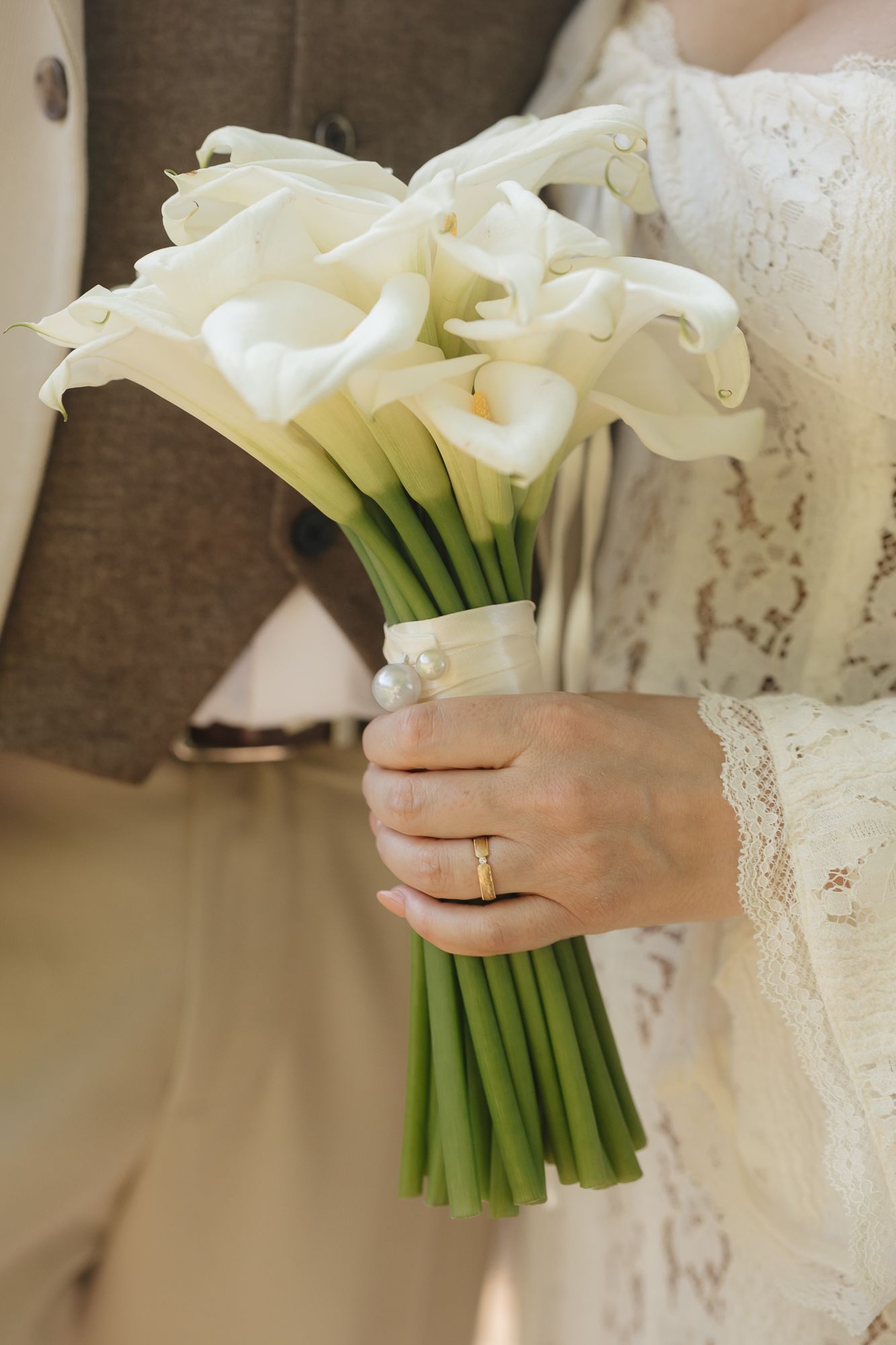A white calla lily bridal bouquet held by hands wearing a delicate lace wedding dress and engagement ring.