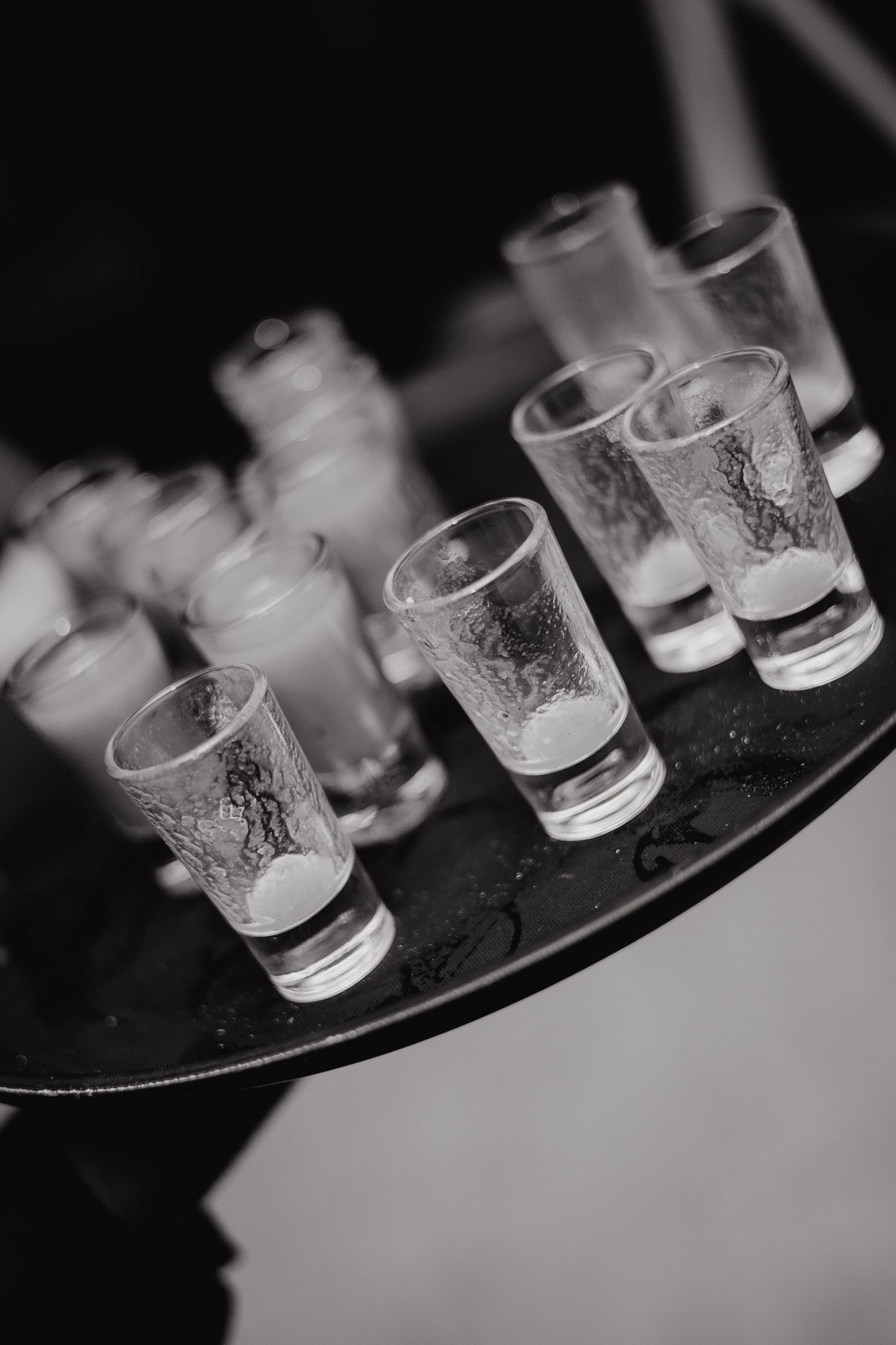 Black and white photo of empty shot glasses arranged on a dark surface in a row, showing motion blur.