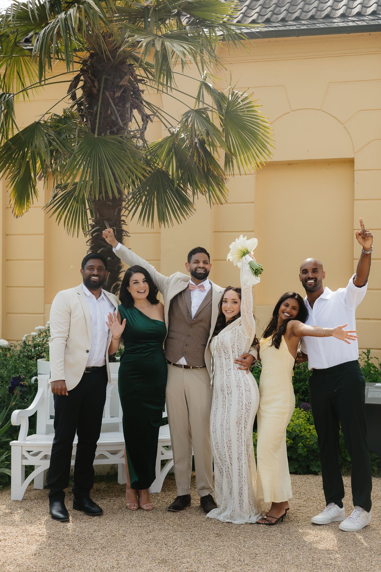 Wedding group photo with guests wearing formal attire against a beige building with palm trees.