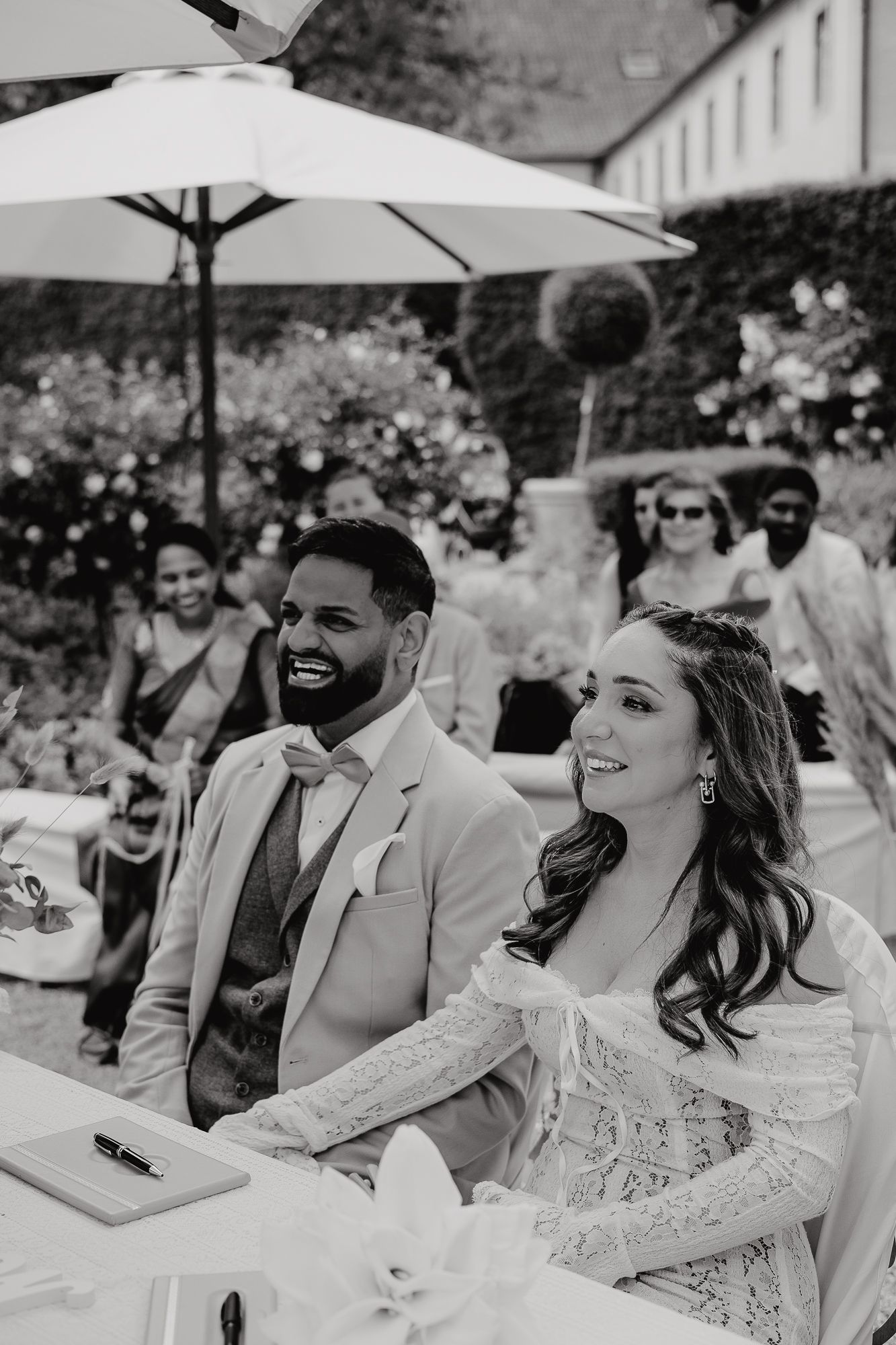 Wedding guests seated outdoors under white umbrellas enjoy ceremony in black and white photography.