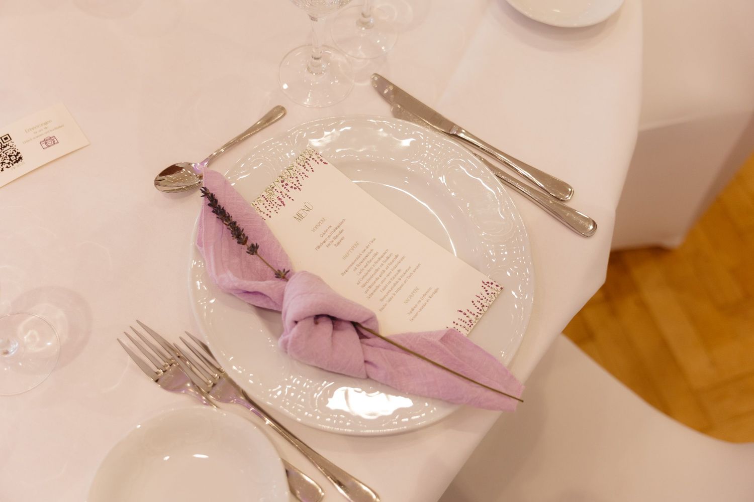 Place setting at an elegant event with a pink napkin, white plate, and formal silverware on a white tablecloth.