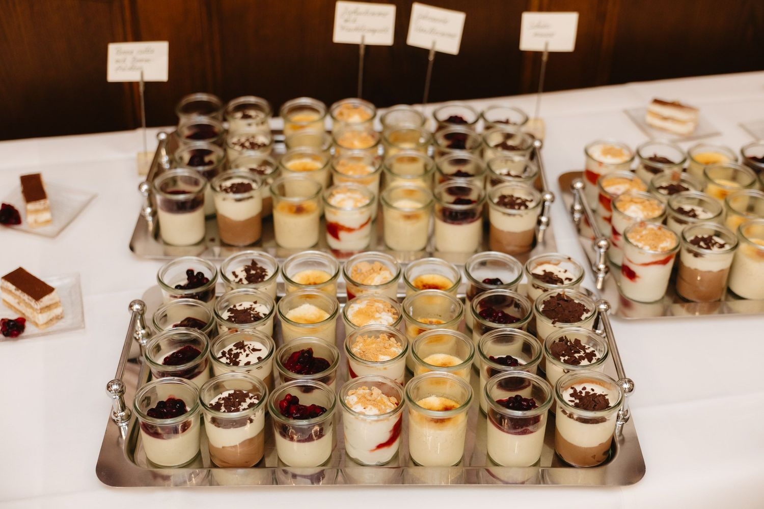 Multiple rows of small glass jars filled with desserts and puddings displayed on a wooden table at a market or event.
