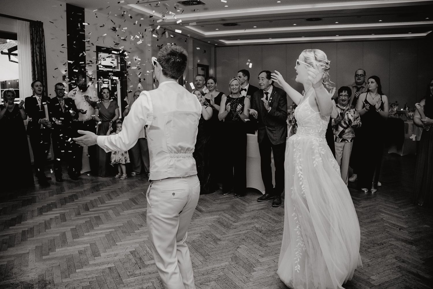 Wedding couple performs their first dance together on a wooden dance floor surrounded by guests at their reception.