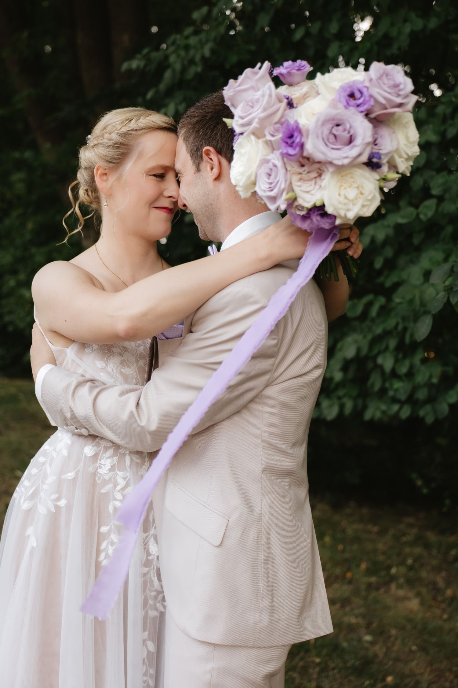 A romantic wedding couple embraces in a garden setting with a purple and white floral bouquet.