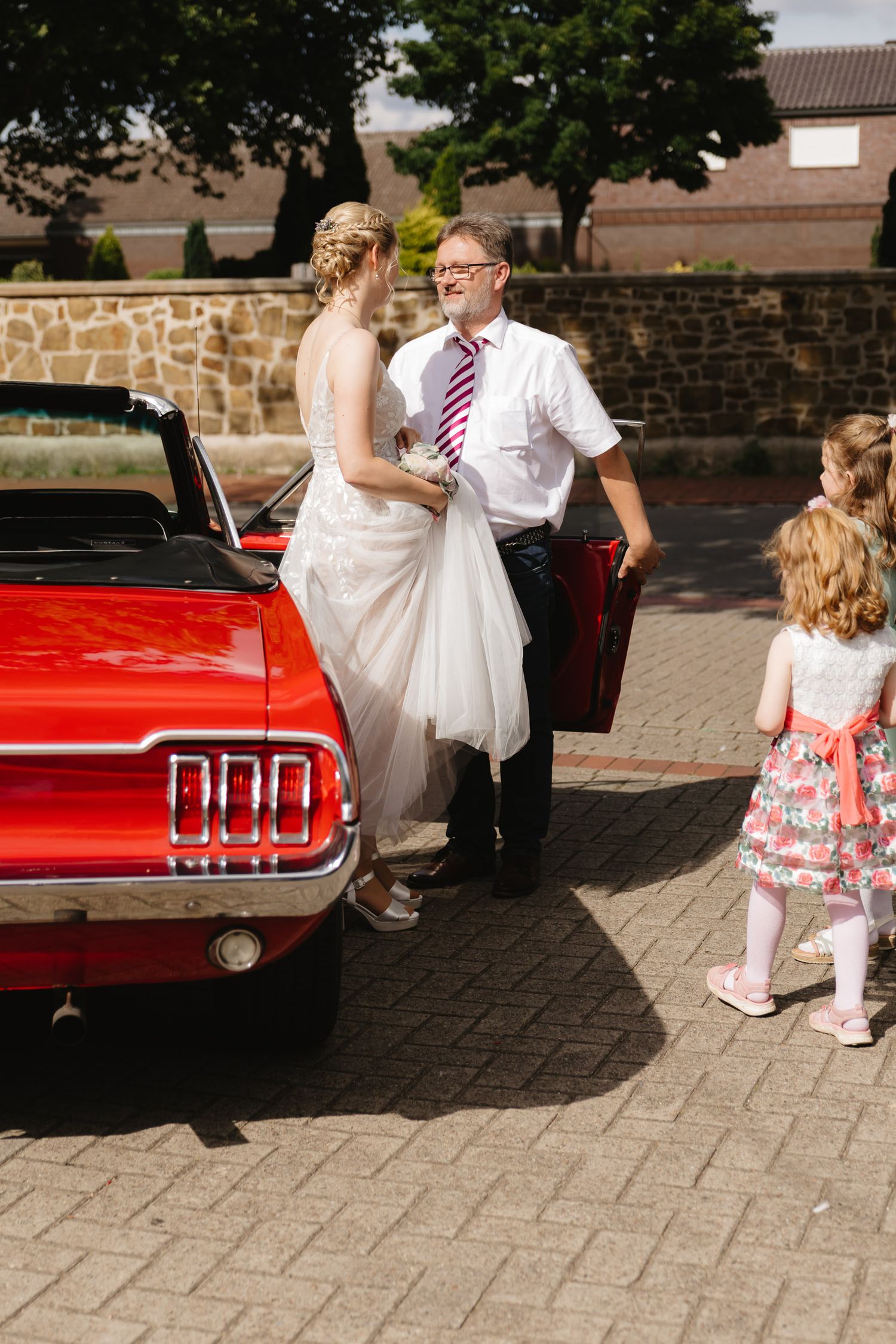 Wedding party members gather around a classic red Ford Mustang convertible on a brick paved courtyard.