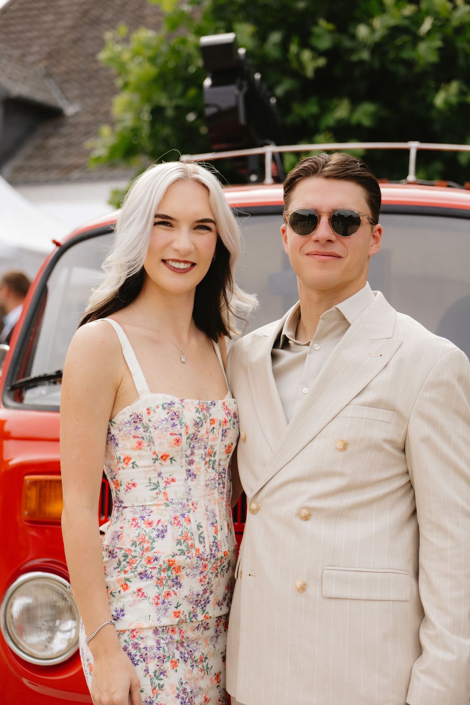 Couple poses next to vintage red car, with bride in floral dress and groom in light colored suit.