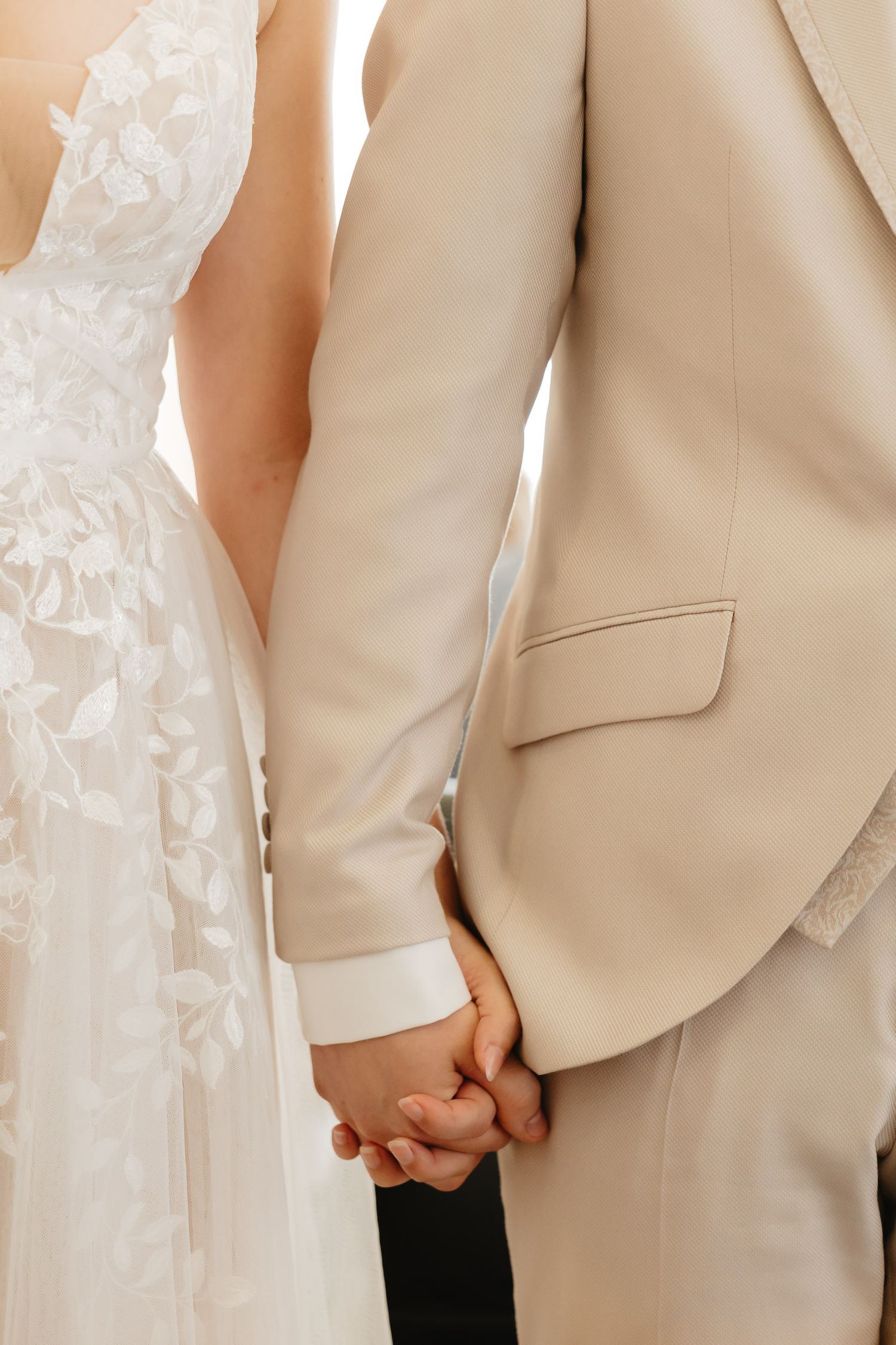 Close-up view of a couple holding hands at a wedding, with bride in white lace dress and groom in beige suit.