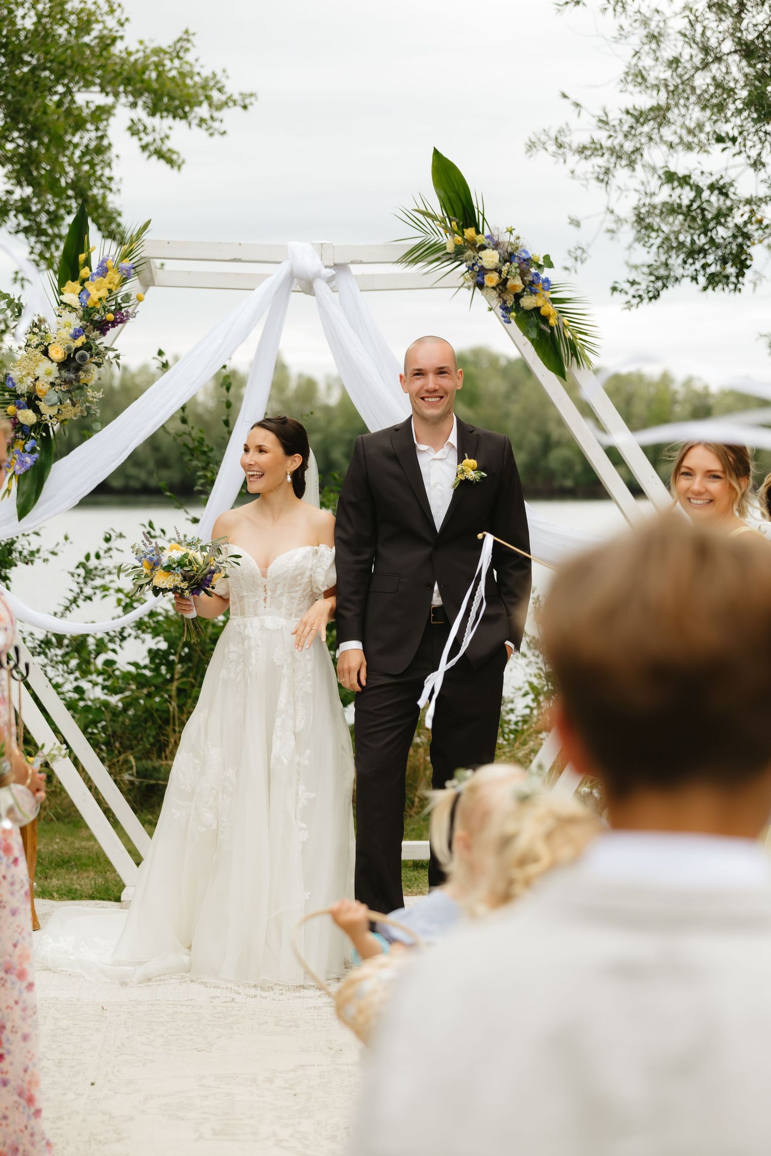 Outdoor wedding ceremony under a white geometric arch decorated with flowers.
