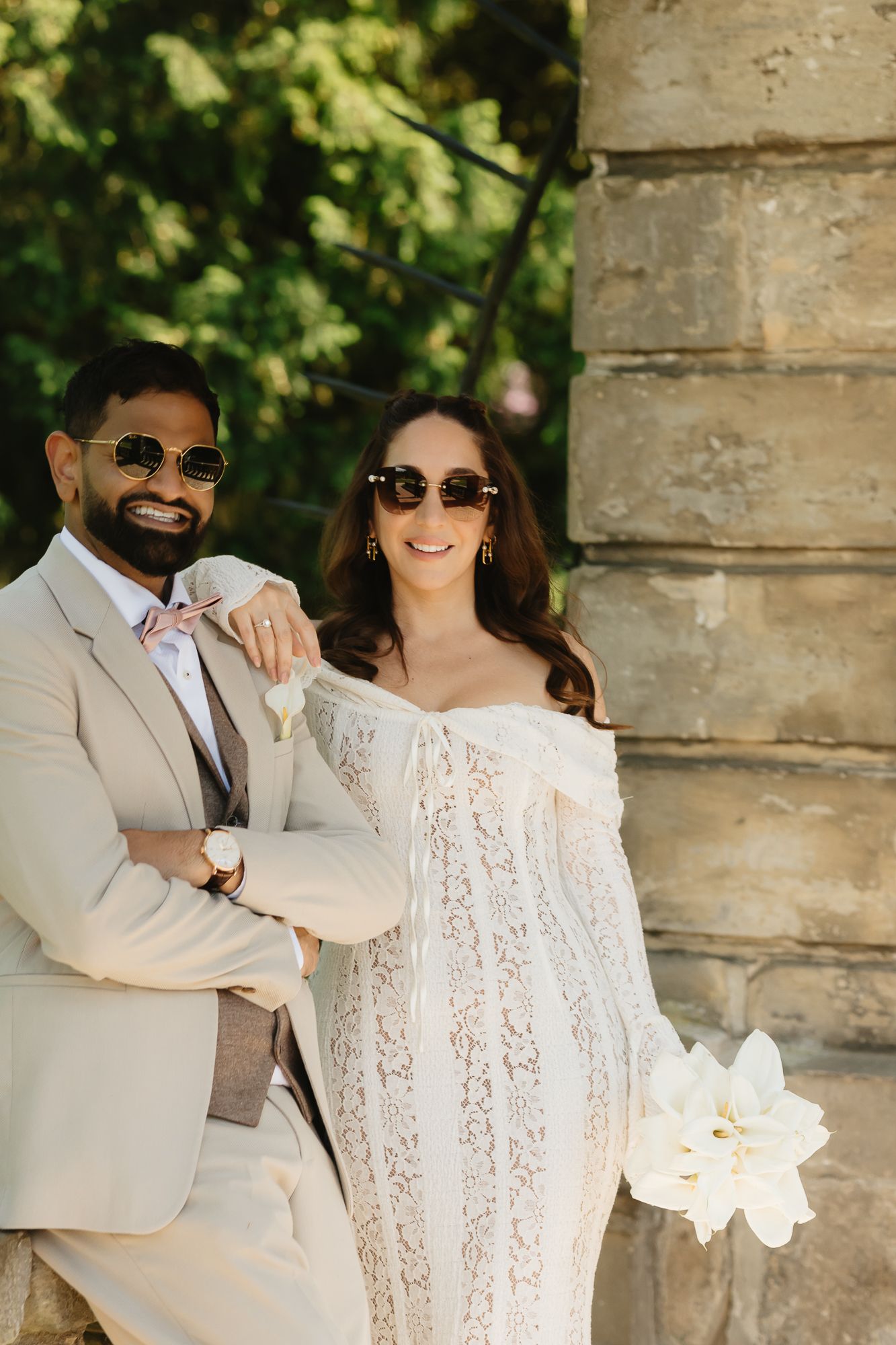 Wedding couple in sunglasses poses playfully with white flowers under stone columns.